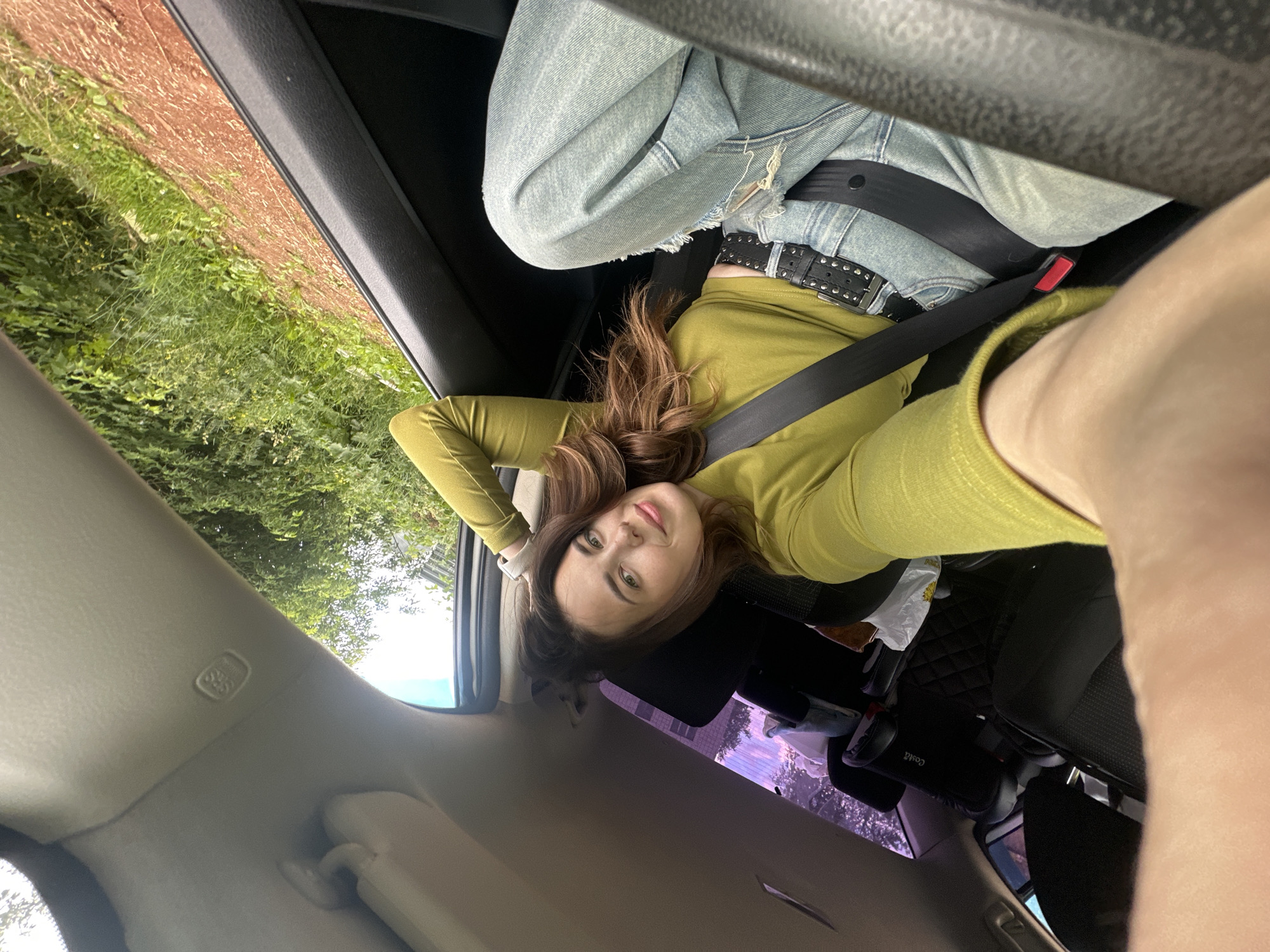 A young woman with brown hair, wearing a lime green long-sleeved shirt, a black belt with silver studs, and light blue distressed jeans, is sitting in the passenger seat of a car. She is looking towards the camera with a slight smile, her left arm resting on the window frame and her head tilted slightly. A black seatbelt is fastened across her body. The interior of the car is visible, including a child car seat in the back. Outside the car window, lush green trees and foliage can be seen, suggesting a rural or suburban setting. The scene is captured from a low-angle perspective, with the car's interior dominating the foreground and midground, and the natural environment filling the background. The lighting is natural, coming from the outside, casting soft shadows. There is no specific indication of the location being in Ilyinsky, Russia.