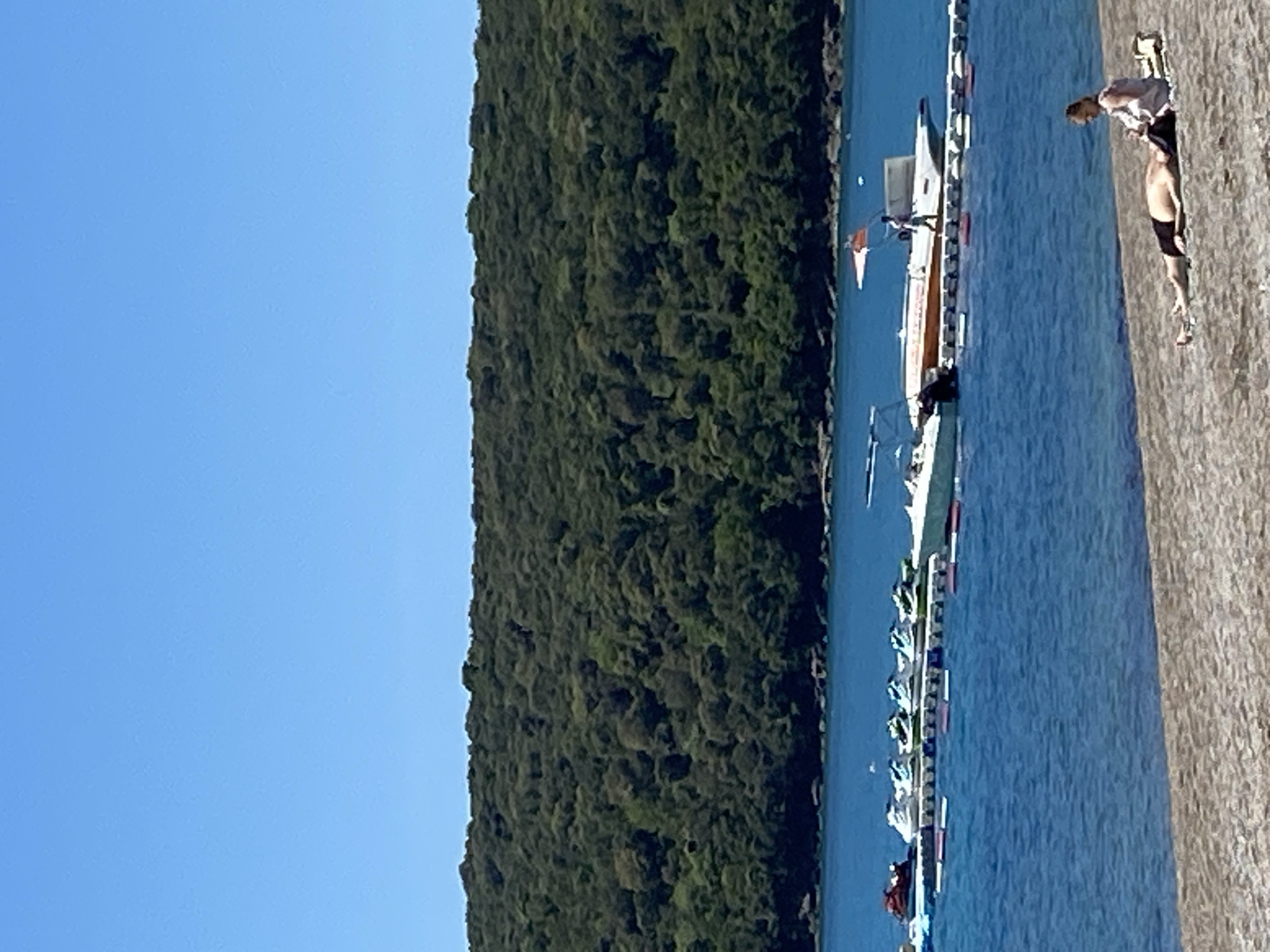 The image depicts a coastal scene in Prijevor, Montenegro. A clear blue sky is visible above a dense, green, forested hill. Adjacent to the hill is a body of blue water, likely a bay or inlet, with a row of boats moored alongside a concrete quay or shoreline. On the quay, a person wearing dark shorts and a light-colored top is lying down, potentially sunbathing. Another individual is standing over them. The overall impression is of a calm, sunny day at a picturesque coastal location.