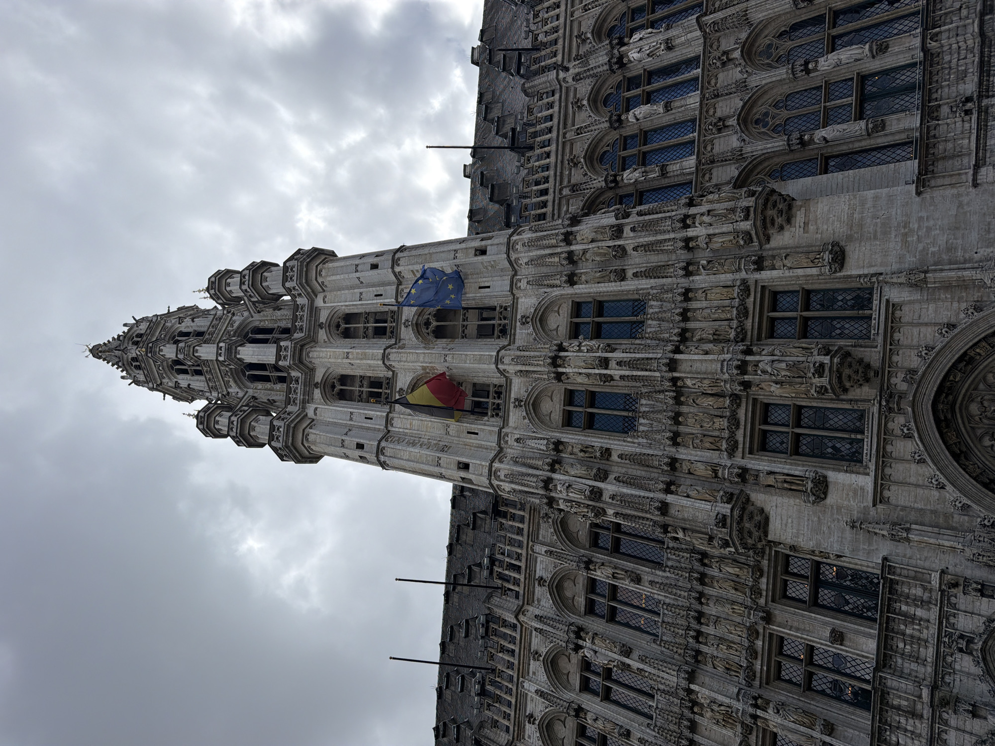 A low-angle, vertical photograph captures a building with intricate architectural details, likely located in the city of Unknown, Unknown. The building's facade is adorned with multiple windows and decorative sculptures. A flag of the European Union and the Belgian flag are prominently displayed on the structure. The sky above is overcast, with a gray, cloudy appearance.