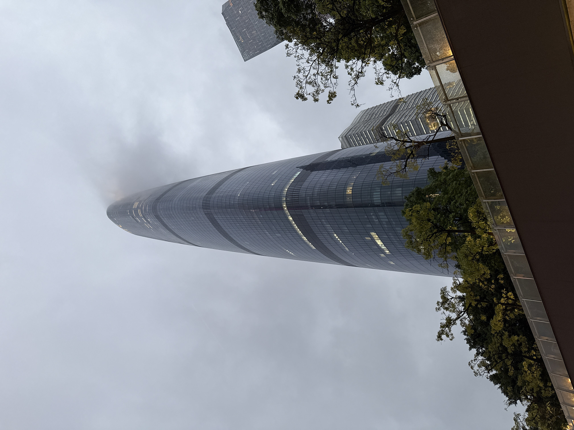 A low-angle shot showcases the Guangzhou CTF Finance Centre skyscraper in Tianhe District, China. The top of the tower is obscured by fog. The sky is overcast and grey. Parts of nearby buildings are visible on either side of the main tower, with some trees partially obscuring the view. The scene is framed by an architectural structure in the foreground, hinting at the viewer's perspective being from an elevated walkway or building.