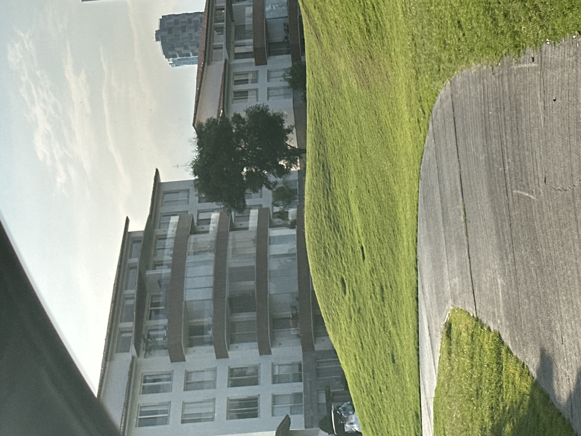 The image depicts a scene in San Bernardino Tlaxcalancingo, Mexico, showing a modern, multi-story residential building with multiple balconies. A well-maintained grassy slope borders a concrete walkway. A tree partly obscures the building. In the background, another building can be seen. No people are clearly visible.