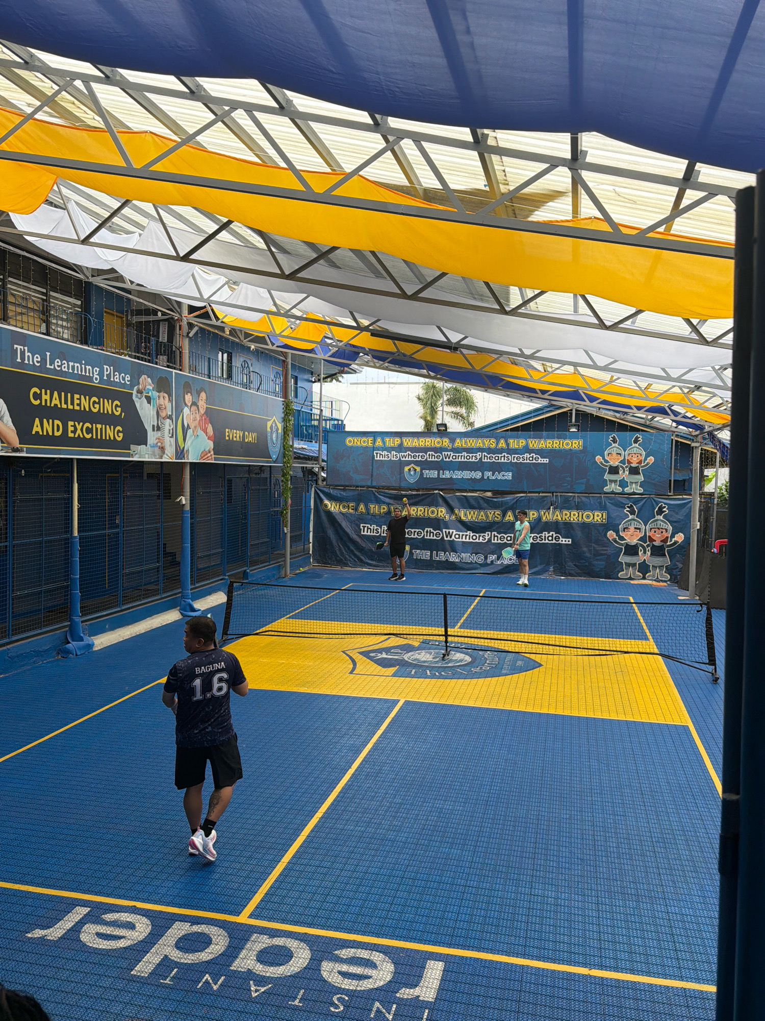 An outdoor pickleball court in Los Baños, Philippines, is captured under a blue and yellow tarp roof supported by a metal frame. The court surface is blue with yellow lines and a yellow central court area. A net divides the court. In the foreground, a man wearing a dark jersey with "BAGUNA 1.6" on the back and black shorts is facing away from the camera, standing on the court. In the background, two men are playing pickleball, one serving with a yellow ball. The court is surrounded by blue fencing and buildings. Banners on the building walls display the text "The Learning Place" and "ONCE A TLP WARRIOR, ALWAYS A TLP WARRIOR!" along with illustrations of children in armor. The overall lighting suggests it is daytime.