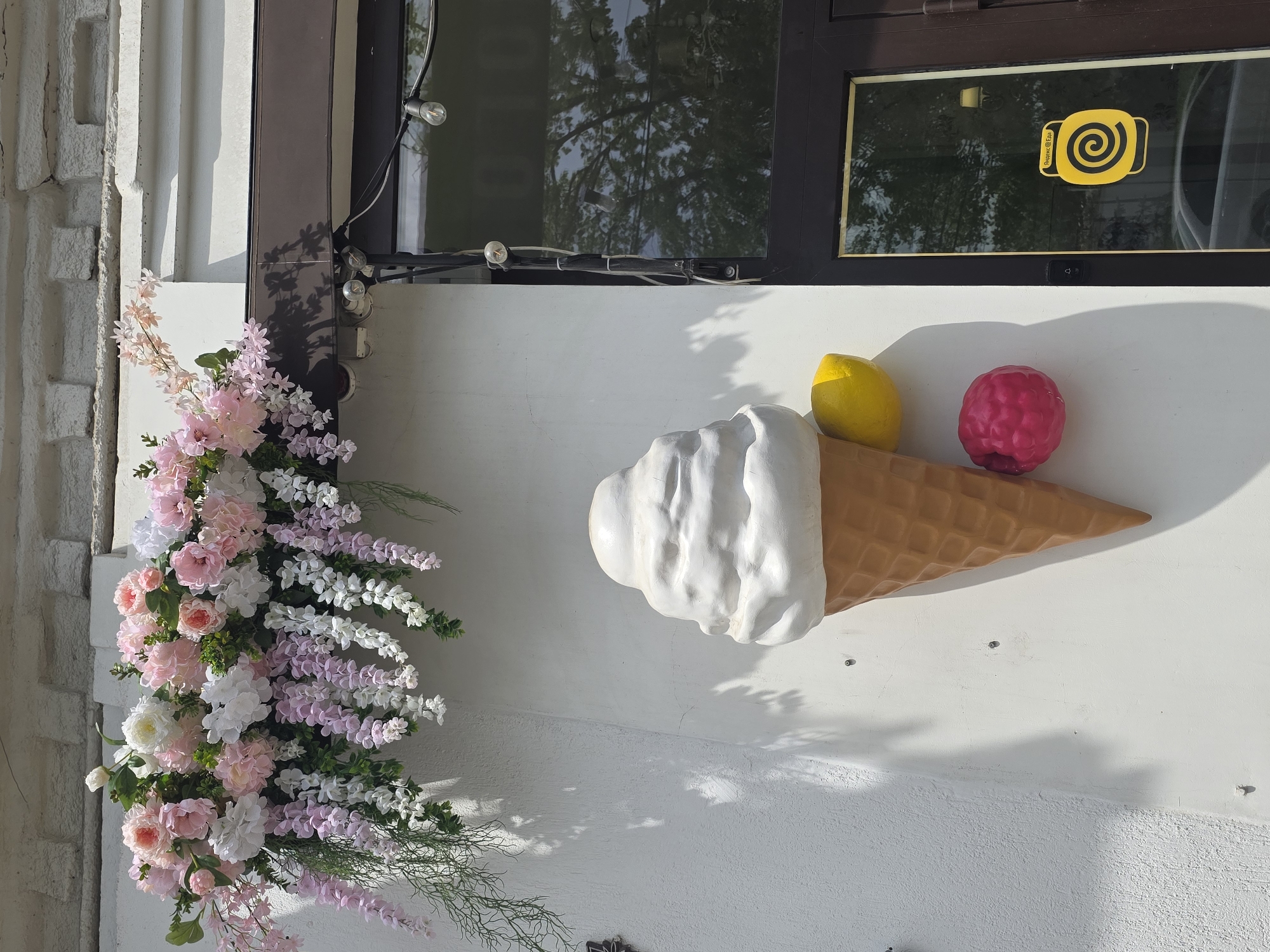 The image shows a building facade. On the left, there is a display of pink and white flowers. To the right, there is a large, decorative ice cream cone mounted on the wall. The ice cream has a white scoop, a yellow lemon-shaped piece, and a red raspberry-shaped piece. Above the ice cream, there is a window with a yellow logo featuring a swirl design and text that includes "Индекс @Еды". Based on this logo and the text visible, the location is likely Krasnodar, Russia.