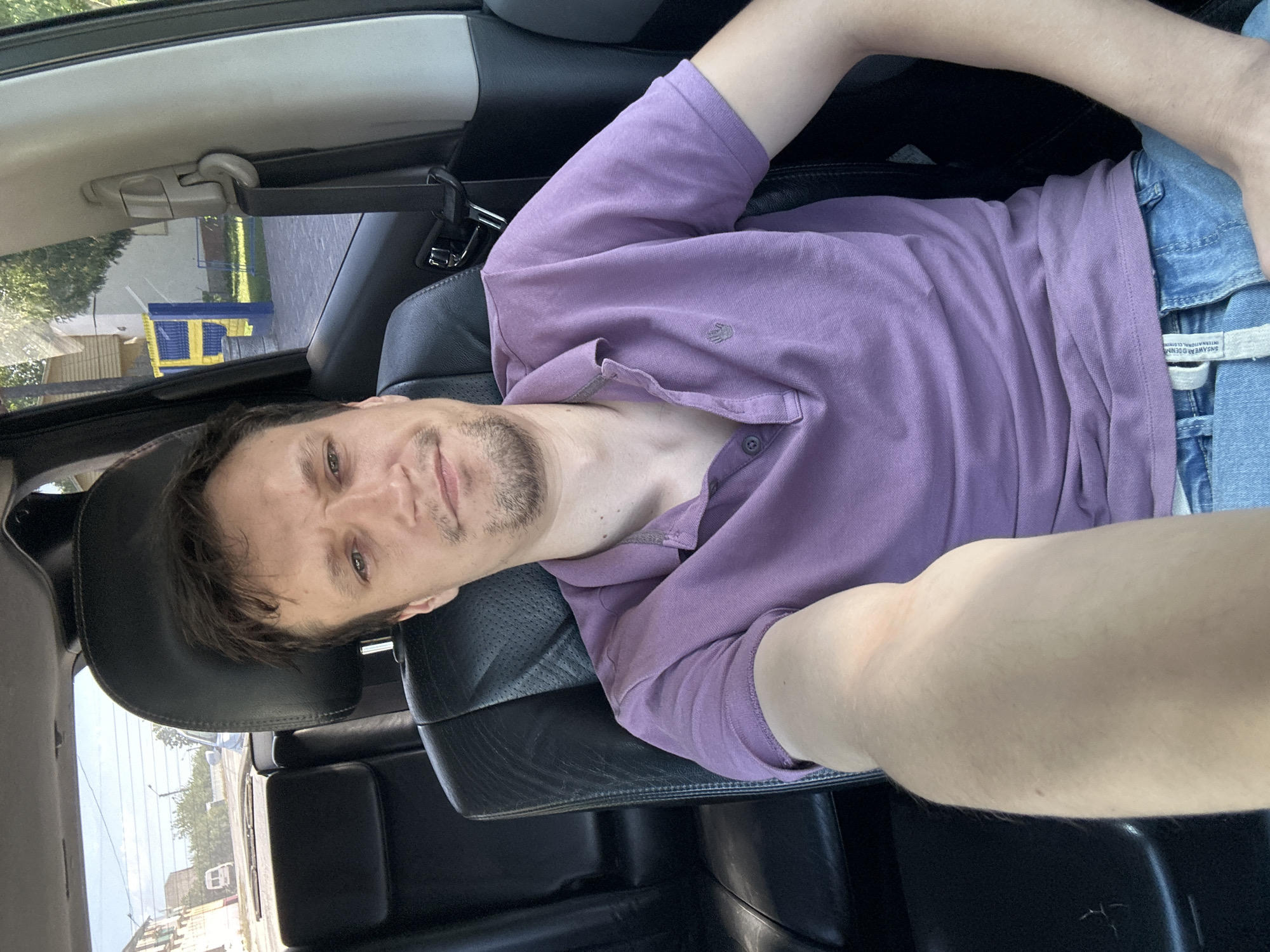 A man with light brown hair and a goatee is sitting in the driver's seat of a car, looking at the camera. He is wearing a purple polo shirt and blue jeans. The car's interior is visible, with black leather seats and a black dashboard. Through the car windows, a street scene can be seen, with buildings, trees, and a white van in the distance. The location appears to be Makiv, Ukraine, based on the context provided. The man's left arm is extended towards the camera, suggesting he is taking a selfie. The overall lighting indicates it is daytime.