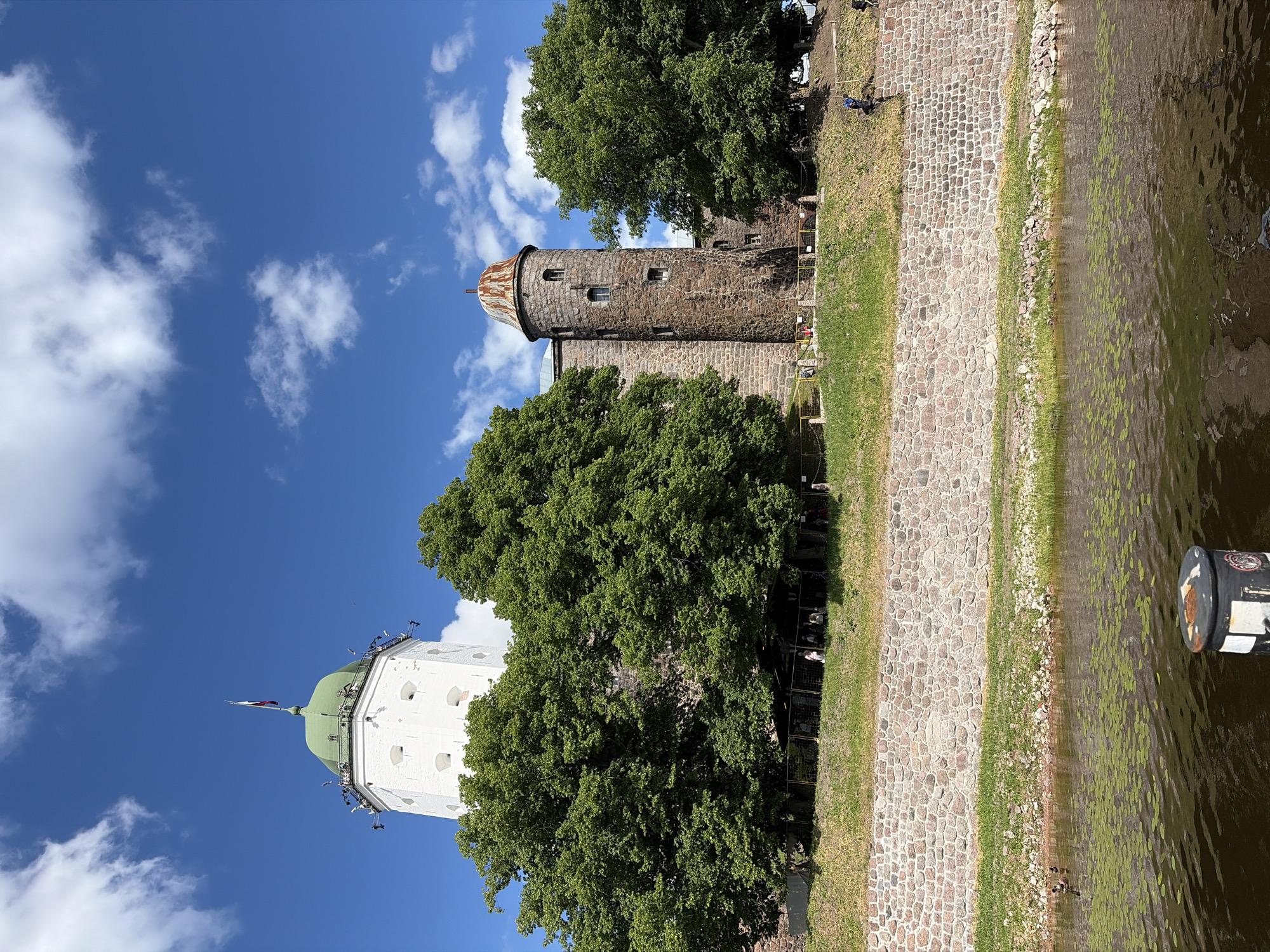 The image shows the Vyborg Castle. The castle has a white tower with a green dome and a Russian flag, and a stone tower with a rusty roof. There are trees around the castle and a river in front of it. The sky is blue with white clouds. There are some people walking on the grass in front of the castle. There are also some ducks swimming in the river. The water in the river is dark brown.