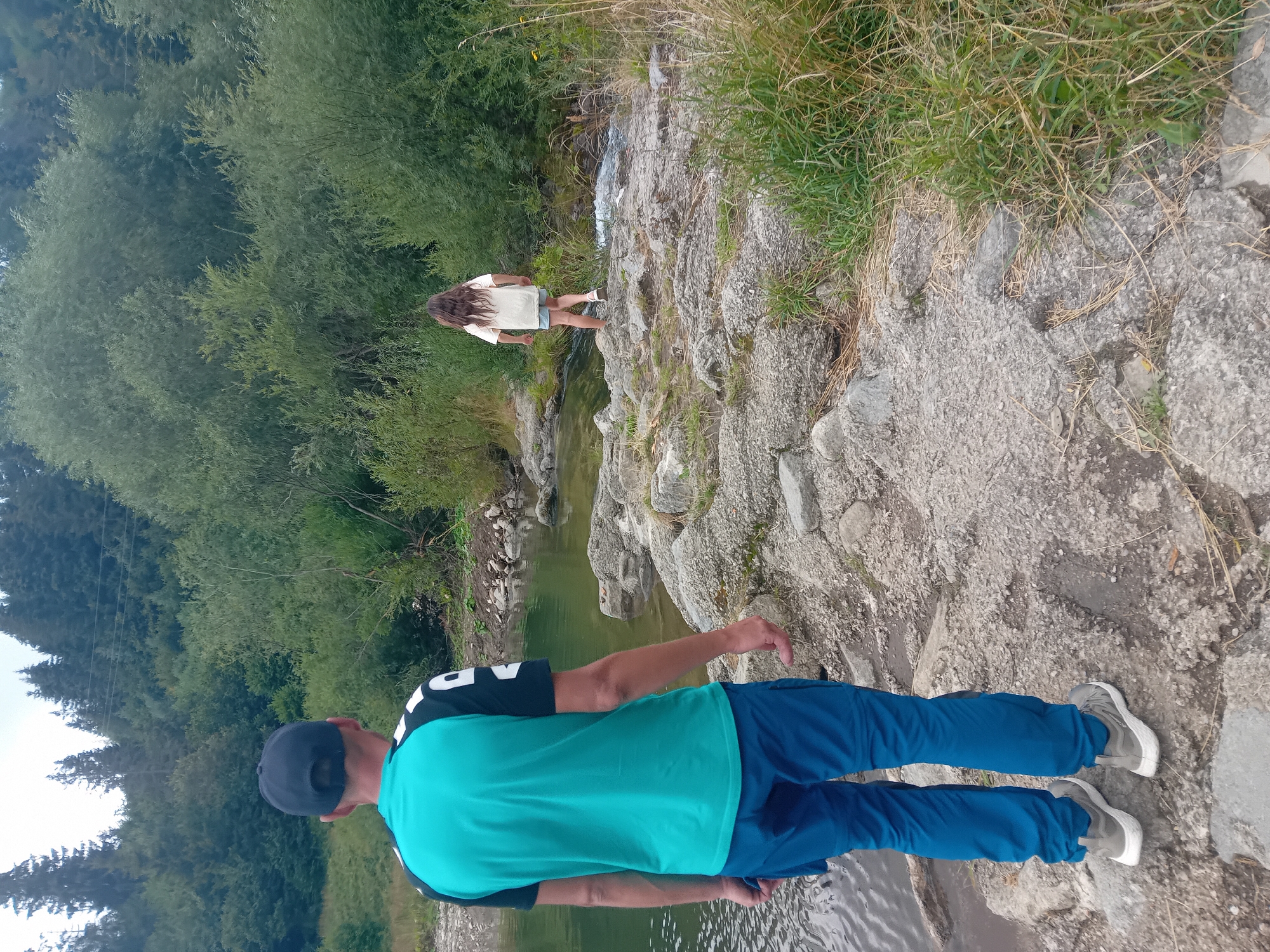 In Habovka, Slovakia, a man wearing a teal and black t-shirt and blue pants stands facing a body of water with a rocky shore. Behind him, a young girl with long brown hair, wearing a light-colored t-shirt and denim shorts, walks on the rocky path towards a small stream. The environment consists of a river or lake, a rocky shoreline, and dense green foliage and trees in the background, suggesting a natural, outdoor setting.