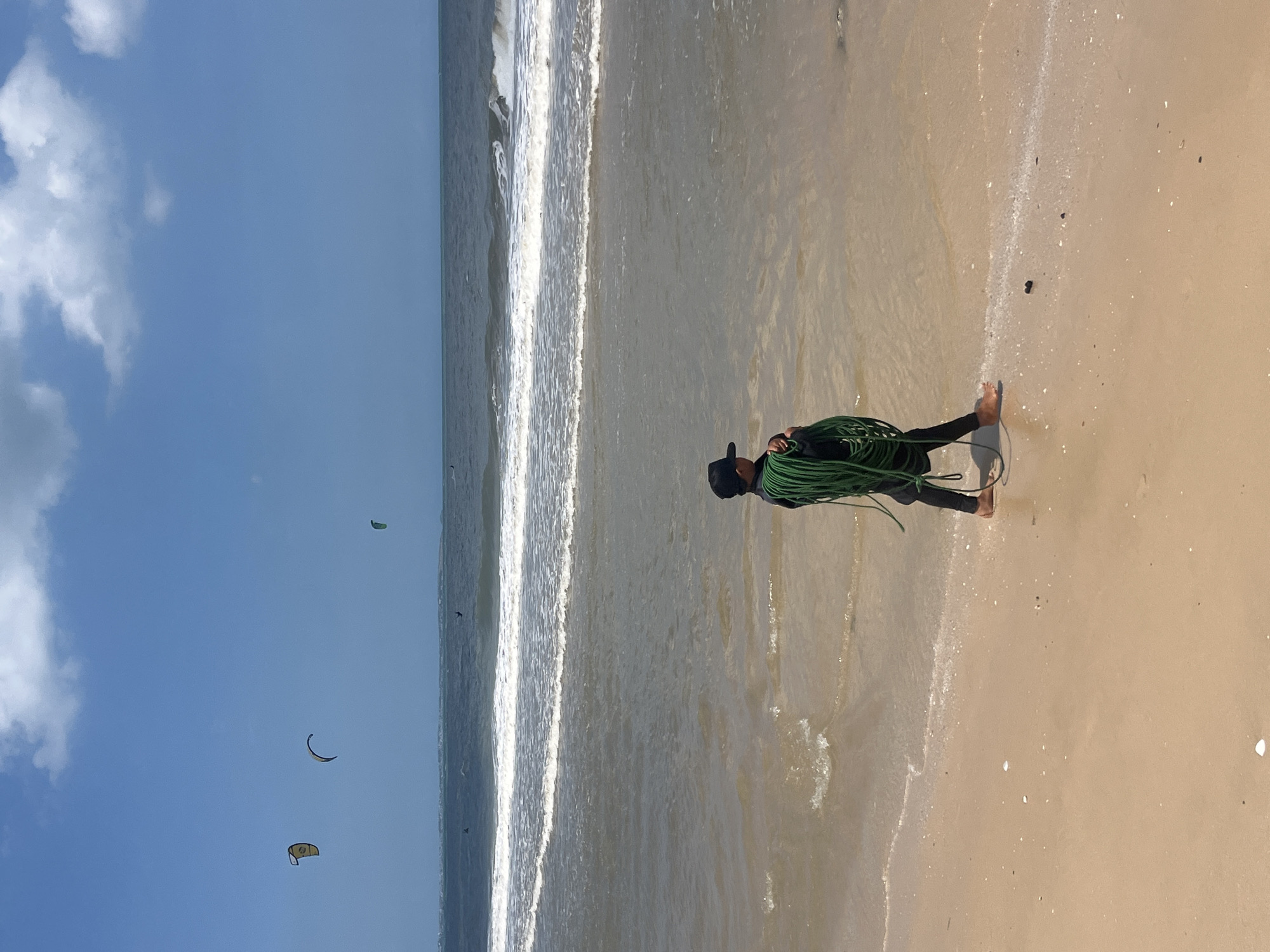 A person wearing a black hat and a dark wetsuit walks along the shoreline of a sandy beach, carrying a coiled green rope over their left shoulder. The person's right leg and foot are visible and bare. The ocean is calm with gentle waves breaking on the shore. Several kites are visible in the sky above the water, indicating a location suitable for watersports like kitesurfing. The sky is blue with scattered white clouds. The image appears to have been taken on a sunny day. The specific location of Phu Thuy Ward, Vietnam, is not discernible from the image itself but is provided as context.