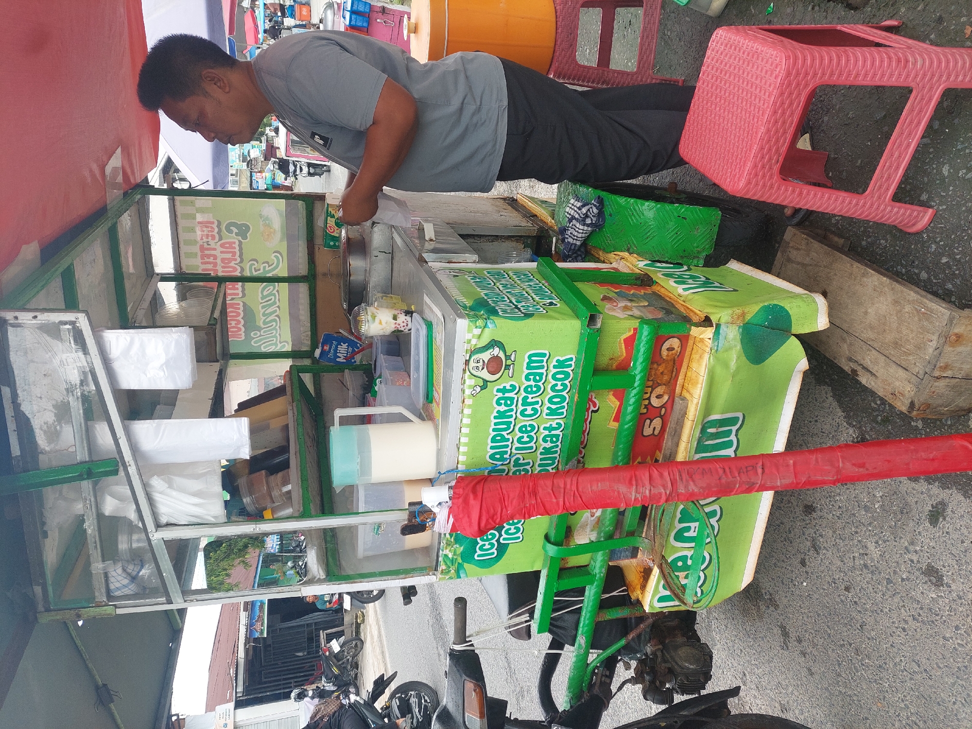 An Indonesian man stands behind a mobile ice cream cart adorned with green and white branding. He is wearing a grey t-shirt and black trousers and appears to be preparing or serving an order. The cart itself is equipped with containers for ingredients, a pitcher of liquid, and a red-wrapped pole. In the background, a street scene unfolds with other vendors, motorcycles, and buildings suggesting a marketplace or busy urban area in the City of Pematang Siantar, Indonesia. The ground is paved with asphalt and there are red plastic stools near the cart.
