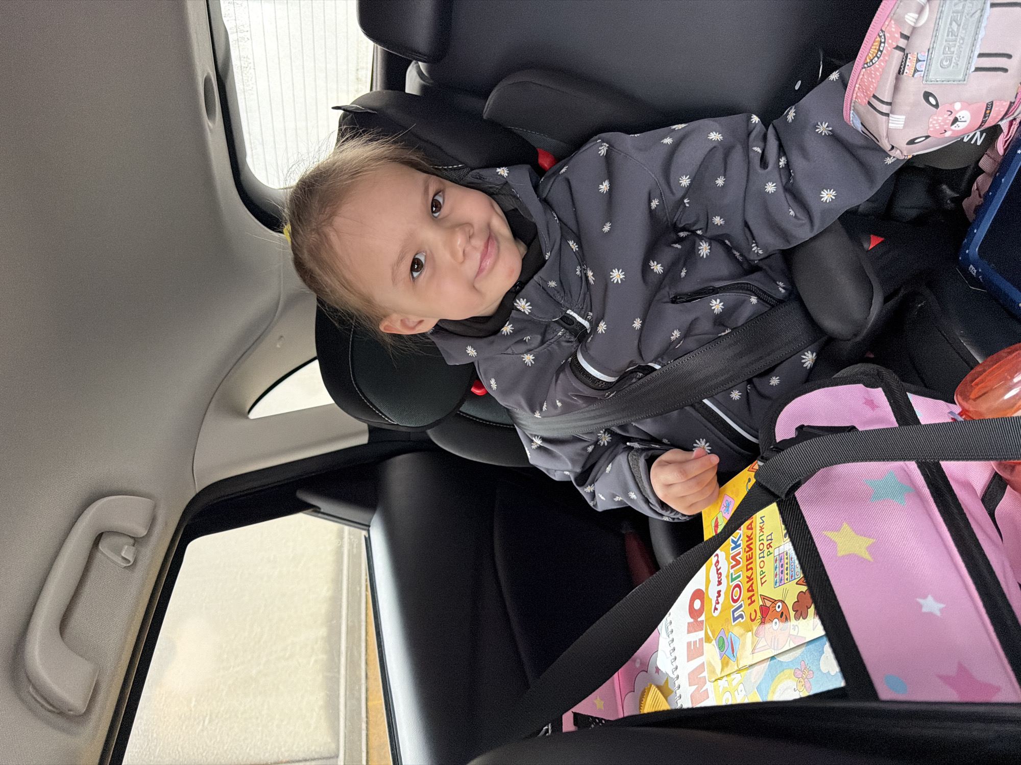 A young child is seated in a car seat within a vehicle. The child, wearing a grey jacket with a daisy pattern, is secured with a seatbelt. Next to the child, there is a pink bag with star decorations, containing books with Russian text, one of which is titled "ЛОГИК" (Logic). The interior of the car is visible, including the car door handle. The location appears to be Izhevsk, Russia, based on the Russian language books.