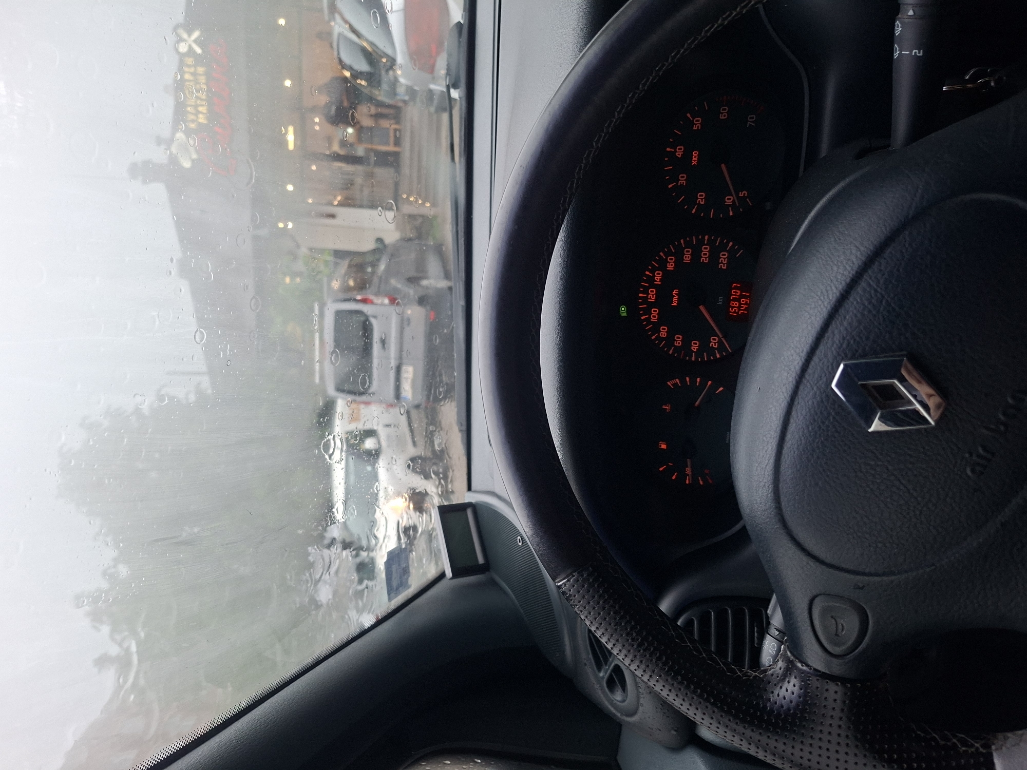 The image captures a driver's perspective from inside a car on a rainy day. The view through the windshield is blurred by numerous water droplets, indicating heavy rain. Outside, a street or parking lot is visible, with several other vehicles, including white vans and cars, appearing indistinctly through the wet glass. In the background, buildings with illuminated signs can be seen. One prominent red sign, partially obscured and distorted by the rain, appears to display text, with OCR suggesting "RYANGAPER MATASUN" on one line and "La Maison Pattaya" on another.
Inside the car, the steering wheel, adorned with a silver Renault logo and "air bag" text, dominates the right side of the frame. The steering wheel features a perforated leather grip section. To the left of the steering wheel, the car's instrument cluster is visible with orange-red illumination. The speedometer reads 0 km/h, with a maximum of 220 km/h. The tachometer also reads 0, with a maximum of 70 x100 RPM. The odometer displays a total mileage of 158707 km and a trip meter reading of 749.1 km. A green indicator light, likely for low beam headlights, is illuminated. The fuel gauge and temperature gauge are also visible, both indicating low levels. A wiper/indicator stalk with settings '1' and '2' is seen to the right of the dashboard. The overall scene suggests the car is either parked or stationary during rainfall.