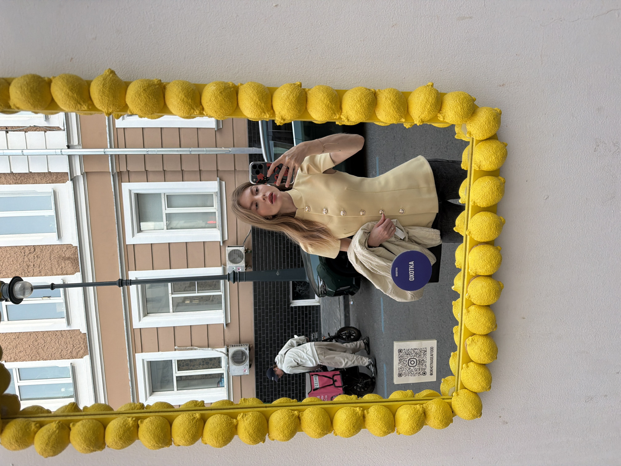 A blonde woman stands in front of a mirror with a lemon-shaped frame and takes a selfie with her phone. Reflected in the mirror is the street scene in front of her. In the background, a brown multi-story building with white-framed windows and a black street lamp extending from the building is visible. A man wearing a white jacket and carrying a pink delivery box is standing on the street. A car is parked near the woman's reflection, and a sign with a QR code and the words "@OKHOTKASEAFOOD" is visible near the bottom edge of the mirror. The mirror also reflects a blue sticker with white lettering that reads "OXOTKA." The background indicates that the woman is located in Moscow, Russia.
