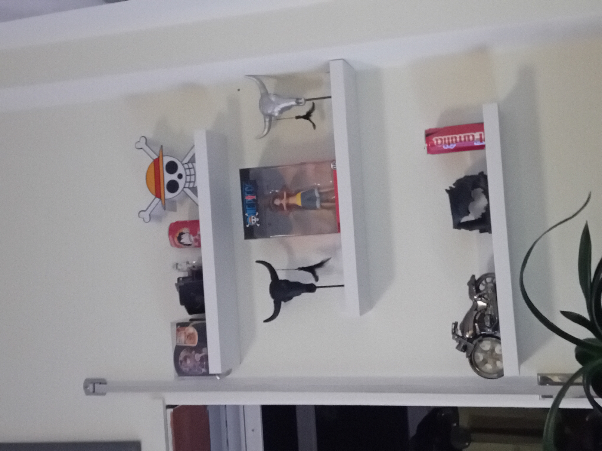 On a white wall are three white shelves displaying various objects. From left to right, the objects are: a One Piece jolly roger symbol, a Pringles can, a black Canon EOS 600D camera, two packs of Nissin Cup Noodles, a silver bull head ornament, a One Piece action figure, a black bull head ornament, a pack of Farukulla candy, a black object that resembles cut paper, and a silver motorcycle ornament. Below the shelves is a window. The environment appears to be a room in a house. The location is Unknown, Unknown.