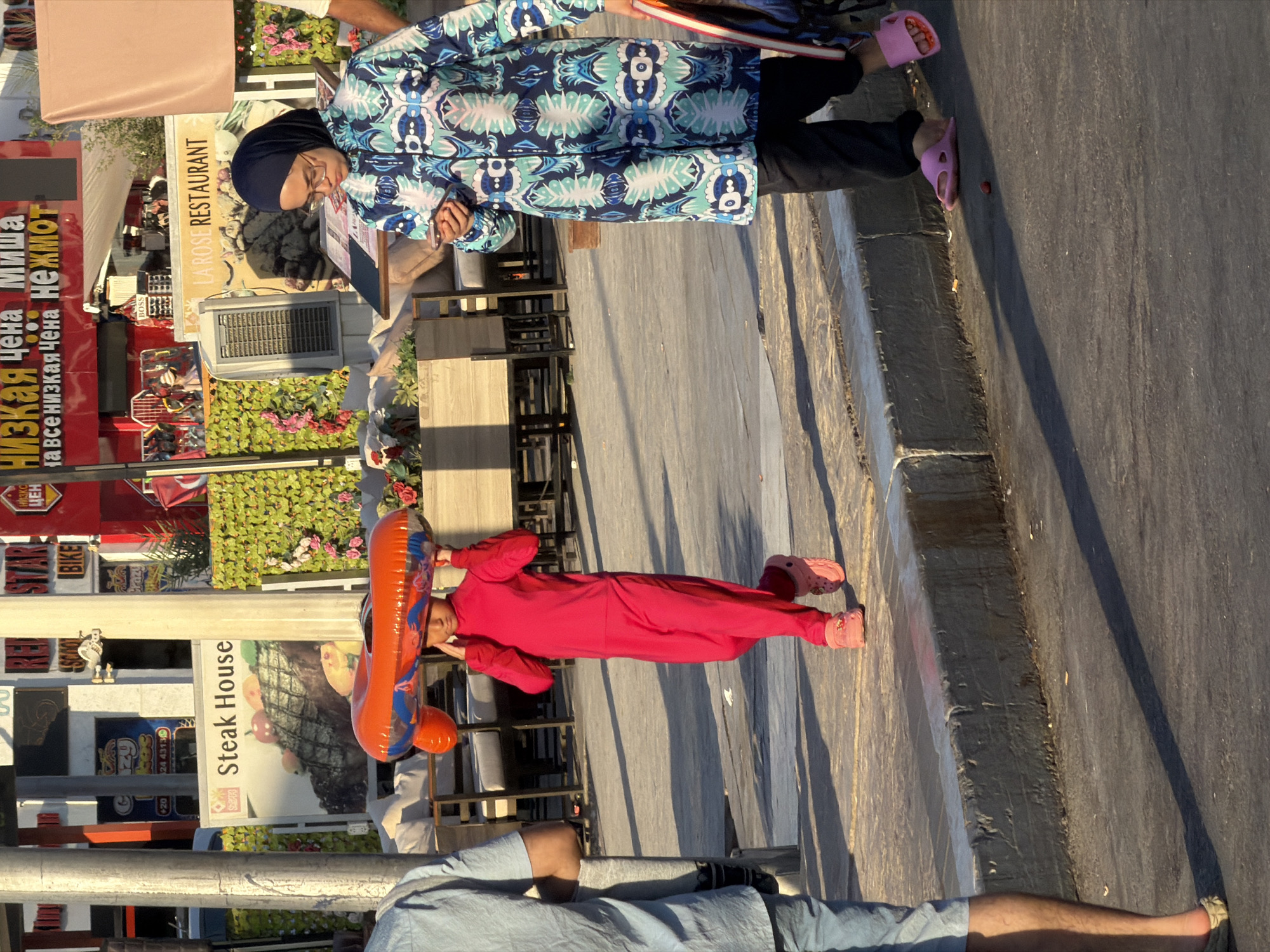 The image depicts a street scene in Sharm El Sheikh, Egypt, likely in a commercial or tourist area. In the foreground, a young person, dressed in a bright pink full-body suit and pink slip-on shoes, walks while balancing an orange inflatable ring on their head. To the right, an adult, wearing a dark headscarf, glasses, and a long patterned blue and green tunic over black pants, is walking and looking down at a smartphone. They are also wearing pink slip-on shoes. To the left, the lower legs of another person are visible, wearing denim shorts and a light-colored sandal.
The background features buildings with signage. One sign clearly reads "Steak House" with an image of a steak. Another sign displays "LA ROSE RESTAURANT". Various plants and decorative elements are visible, including green hedges and potted plants. The ground is paved with concrete and asphalt, with a raised concrete curb separating the sidewalk from the street. The lighting suggests it is daytime, with shadows indicating the sun is present. Several poles are visible on the left side of the image.