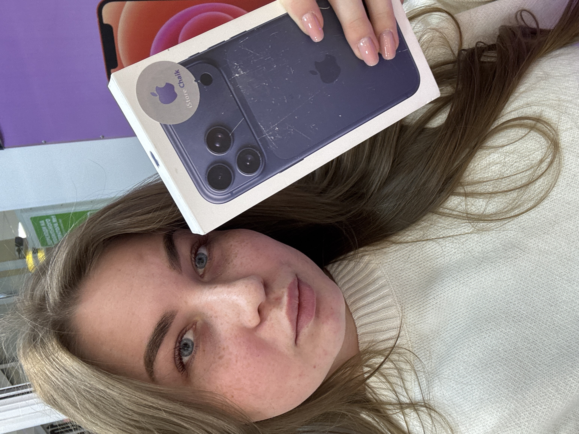 A young woman lies on her side, wearing a white sweater. She holds a box for a blue iPhone. The box has a sticker with the text "iStore Chaikovsky" and an Apple logo, indicating the phone was purchased from a store in Chaikovsky, Russia. The phone on the box appears scratched. The woman has light skin, freckles, light brown hair, and blue eyes.