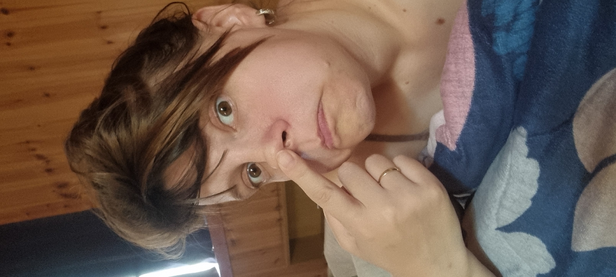 A middle-aged woman with light skin and brown hair is pointing at her nose. She is wearing a gold ring on her left hand. A blue and pink patterned blanket is draped over her chest. The background includes a wooden wall and flooring. Location cannot be determined from the image.