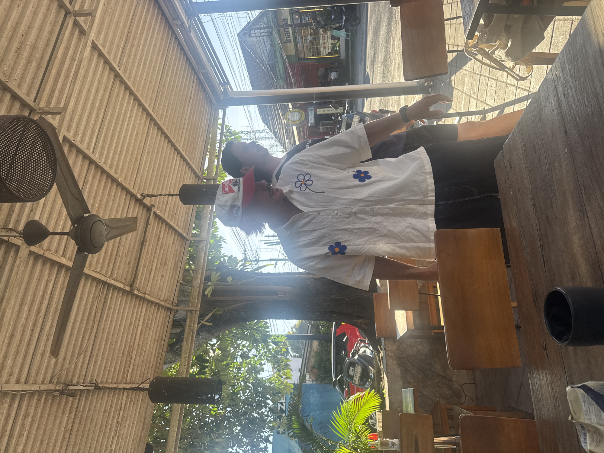Two men sit at a wooden table under a corrugated roof in Kuta Selatan, Indonesia. One man wears a white shirt with flower appliques and a white and red cap. A ceiling fan is visible above. Beyond the immediate structure, lush greenery and street scenes with shops and motorcycles can be observed.