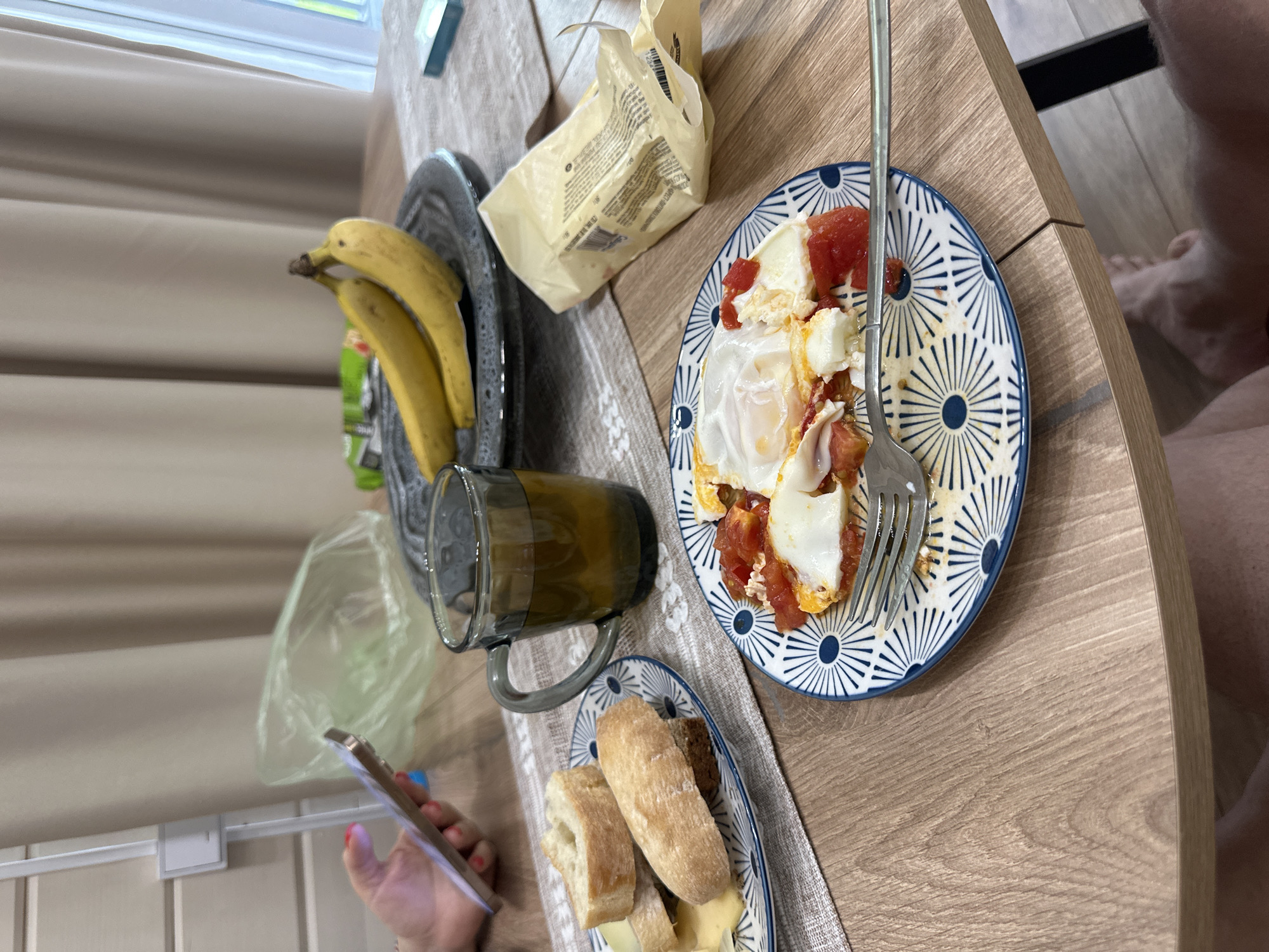 A dining table is set with a breakfast spread. A plate with two fried eggs and sliced tomatoes is in the center. To the left, there is a plate of bananas and a package of butter. A mug filled with a brown liquid is next to a small plate with slices of bread and cheese. A hand holding a smartphone is in the lower left corner. The backdrop includes a curtain and part of a window. A person's leg is visible in the upper right corner. The location is not apparent, and there is no verifiable evidence that the image was taken in the city of Buzhaninovo, Russia.