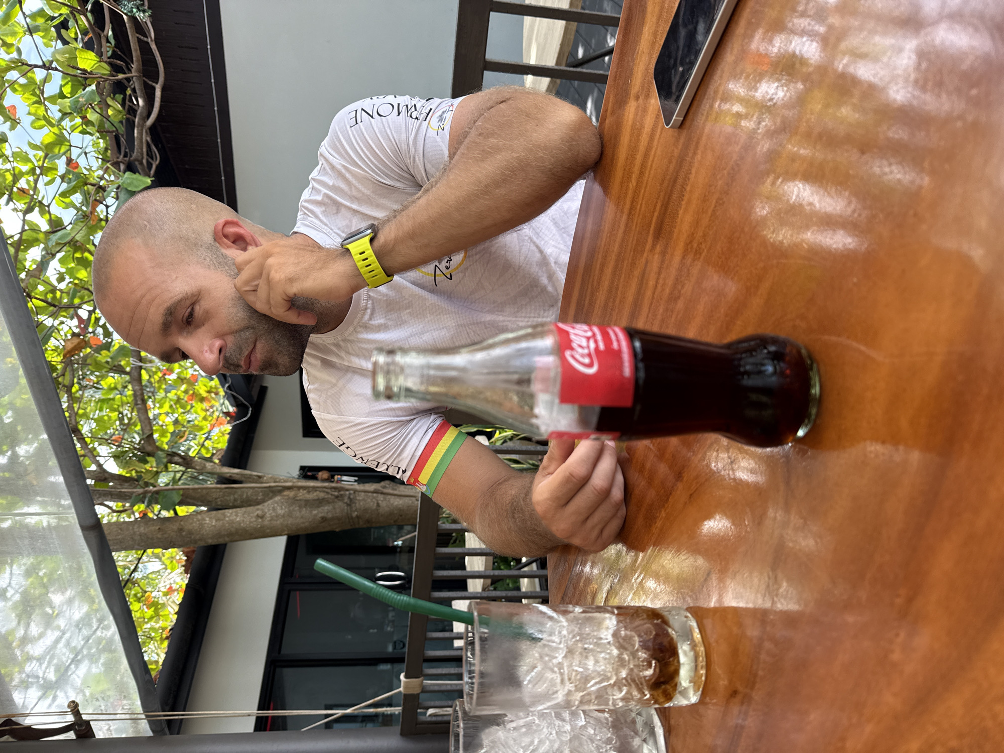 A man with a shaved head is sitting at a wooden table outside. He is wearing a white t-shirt with yellow and red stripes on his left arm and a yellow smartwatch. He is looking to his right, with his right hand resting on his chin. In front of him on the table is a bottle of Coca-Cola, a glass with ice and a green straw, and a smartphone. The table is polished and reflects the light. In the background, there is a tree with green leaves and a building with white walls and dark windows. The scene is well-lit and appears to be daytime. The location is likely a restaurant or cafe in the Ko Samui District of Thailand, suggested by the tropical foliage and architecture.