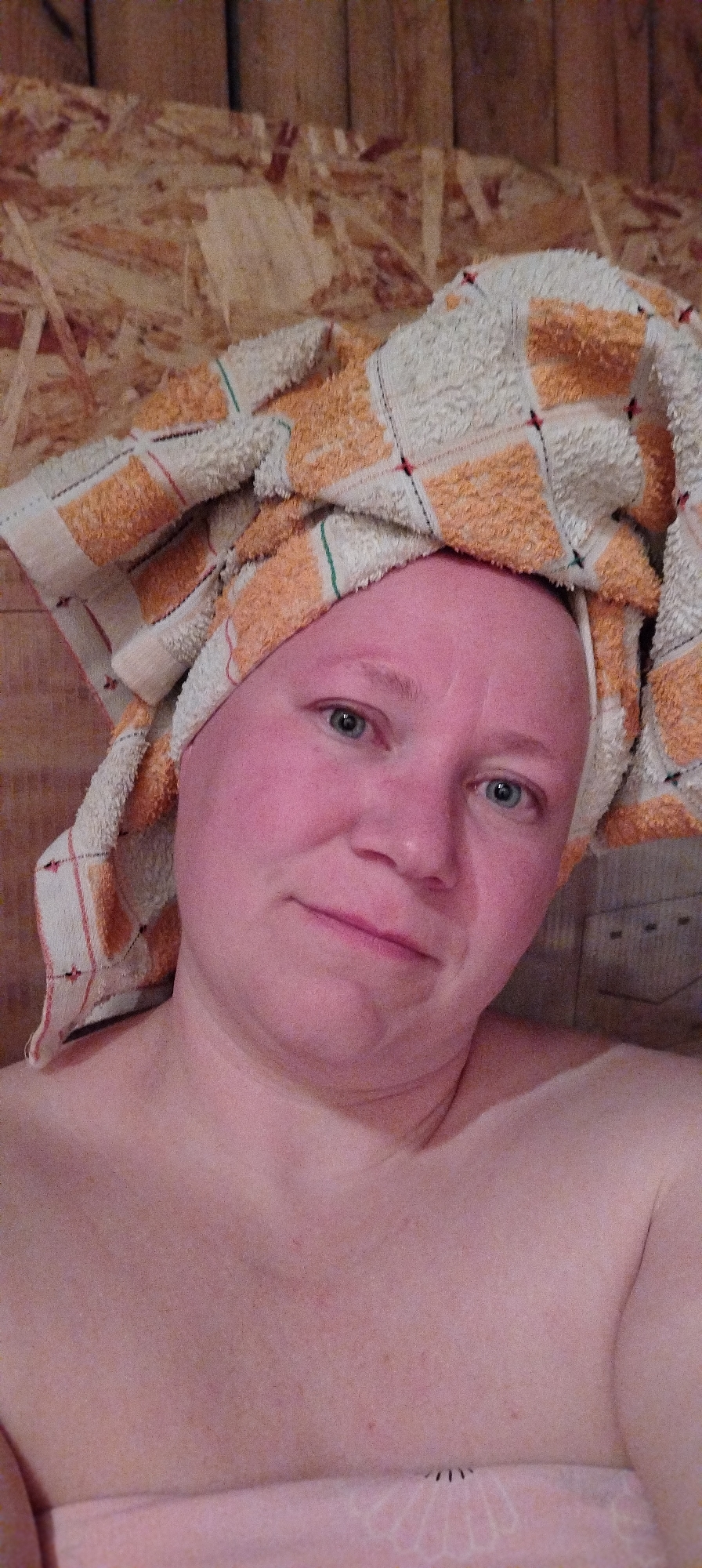 A close-up shot of a fair-skinned person with a towel wrapped around their head, likely taken indoors. The person has blue eyes and a slight smile. The towel is patterned with white and orange squares. The background consists of wood paneling. The image was taken in Зипуново, Russia.
