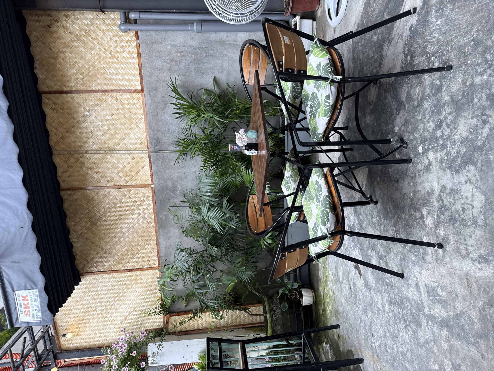 The image depicts an outdoor cafe setting in what appears to be Đà Nẵng, Vietnam. A small, dark wooden table stands between two wooden chairs with black metal frames. The chairs are adorned with white and green patterned cushions. Behind the table is a lush potted plant. The backdrop consists of a concrete wall, partially covered with a woven bamboo panel. To the left, a black awning provides shade. The ground is concrete. A fan is visible on the wall. On the table, there are flowers in a vase and salt and pepper shakers. Another plant and a small, metallic structure sit on the ground. A sign with Vietnamese writing can be partially seen to the left under the awning. The scene is quiet and devoid of people.