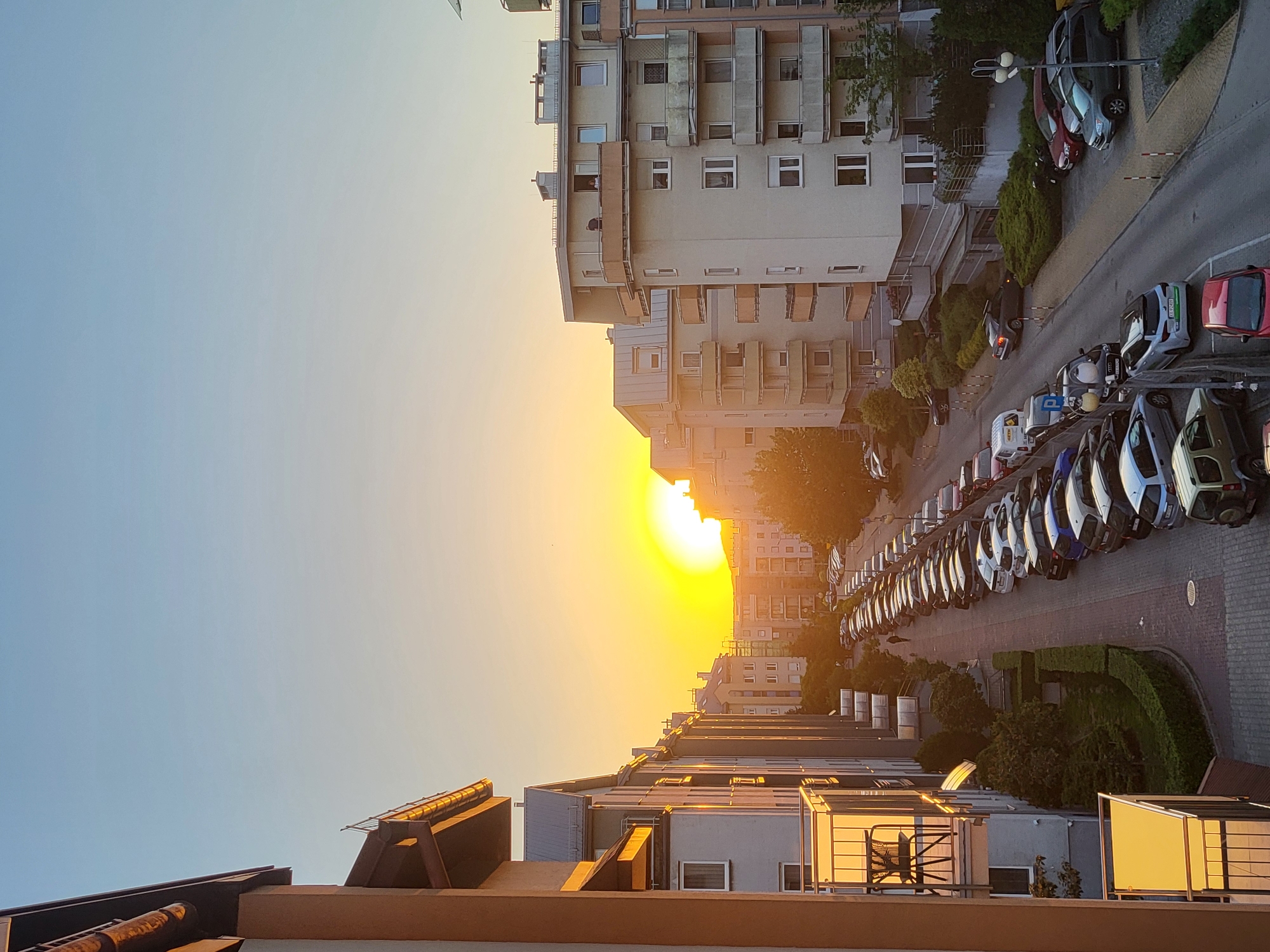 The image depicts an urban scene at sunset in an unknown city. The perspective is from an elevated position, looking down on a street lined with parked cars, mostly white. Buildings are visible on both sides of the street, typical of residential areas. The setting sun is positioned in the middle of the frame, casting a warm, golden glow over the scene. The sky transitions from light blue to orange near the horizon, indicating either sunrise or sunset. Rooftops of buildings are visible in the foreground. There are no people visible.