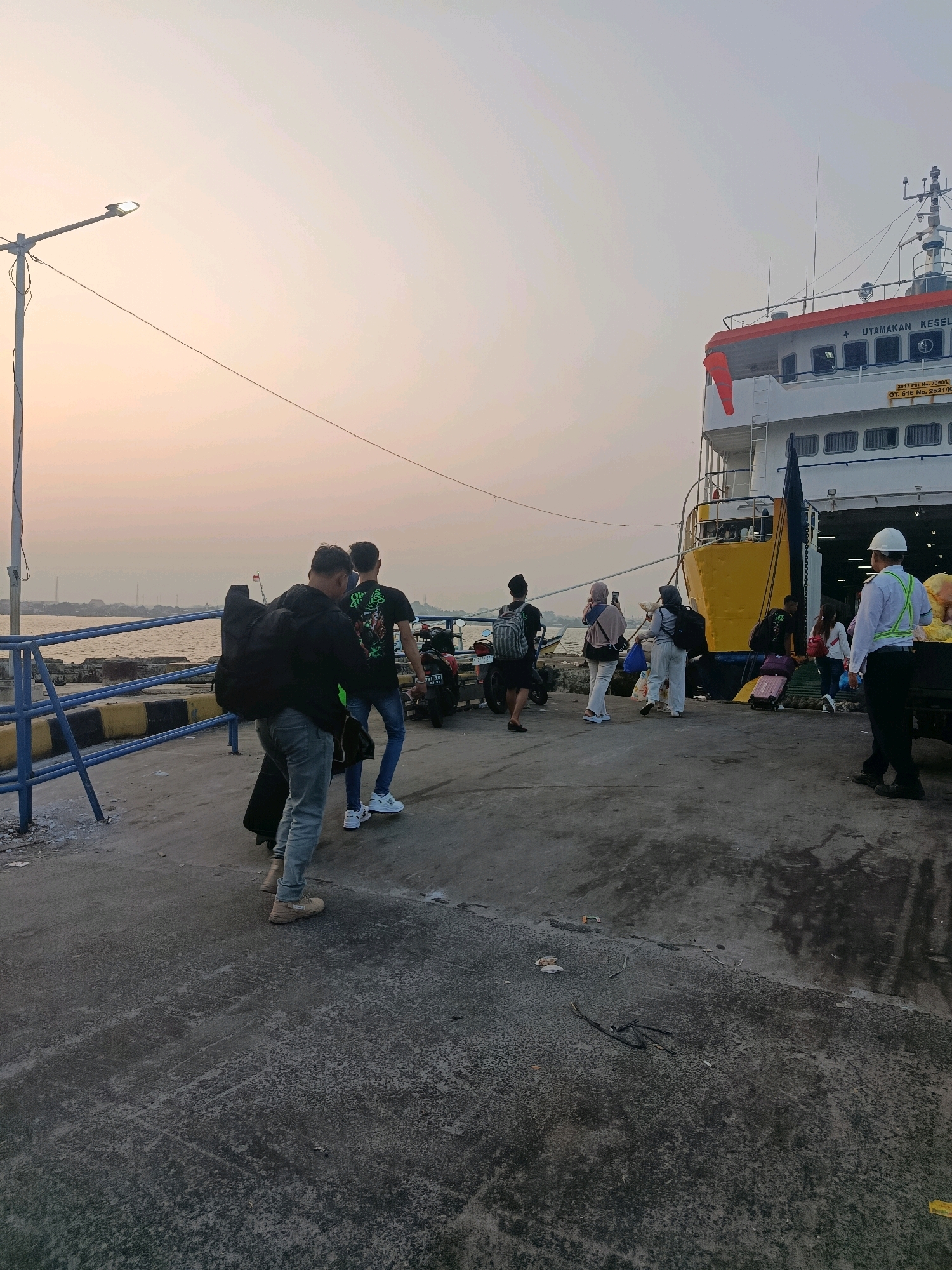The image depicts a busy ferry terminal at sunrise or sunset. In the foreground, two men are walking away from the camera, one carrying a large black bag over his shoulder. The man in front wears a black jacket and grey jeans, while the man behind him is in a black t-shirt and blue jeans. To the right, a yellow and white ferry is docked, with several people seen boarding or disembarking. One person, wearing a white shirt and a reflective vest, appears to be directing traffic or overseeing operations near a truck. Several motorcycles are parked on the pier, and various pieces of luggage are scattered around. The environment is a concrete pier with a metal railing separating it from the water. The sky is a soft gradient of orange and pale blue, suggesting early morning or late evening light. The text on the ferry reads "UTAMAKAN KESELAMATAN". This scene is likely at the Kartini Port in Jepara, Indonesia, which serves as a major hub for inter-island travel.
