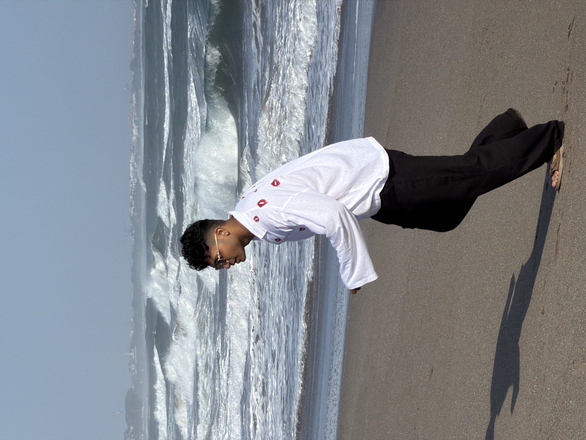 A young man with dark curly hair and sunglasses walks along a sandy beach. He is wearing a long-sleeved white shirt with red kiss prints and black pants. The ocean behind him is rough with large waves breaking. In the distance, a hazy cityscape is visible. The overall scene appears to be in the city of North Kuta, Indonesia.