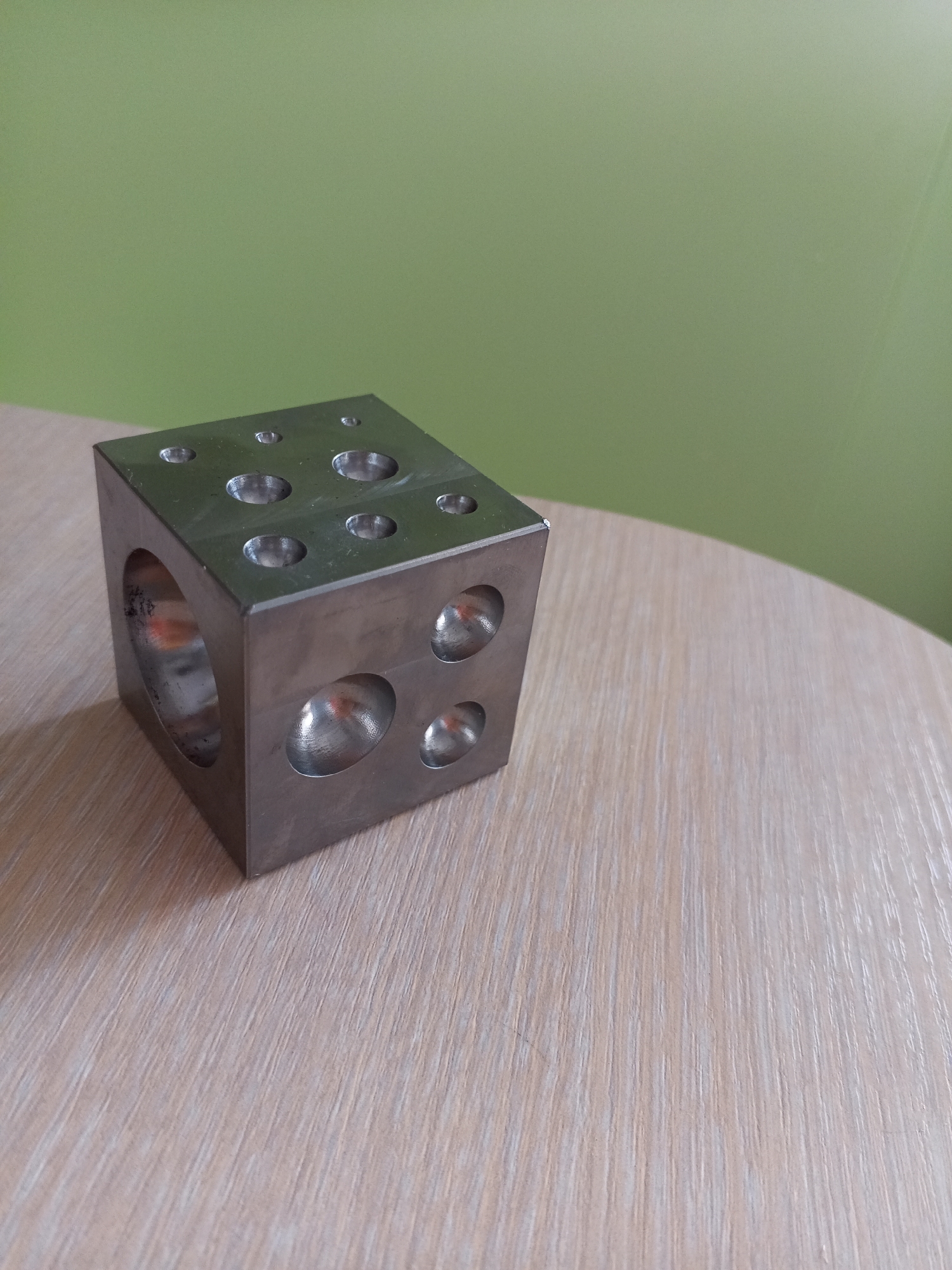 A close-up, eye-level, medium shot captures a metallic cube with multiple hemispherical indentations on its surfaces, resting on a light-colored wooden table. The cube is positioned on the left side of the frame, with its top and side surfaces visible. The indentations vary in size and depth. The background features a solid, light green wall. The scene is indoors, with a light source casting reflections on the metallic surface of the cube and the wooden table. The image does not provide any information that could place its location in Vilyuysk, Russia, or any other specific city. There are no people, actions, or interactions depicted.