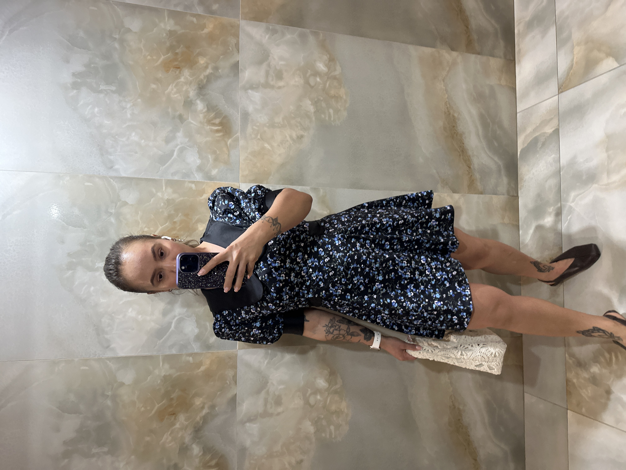 A woman stands in front of a tiled wall, taking a selfie with her phone. She is wearing a short, dark floral print dress with puff sleeves and a black collar, and brown slip-on shoes. She is holding a white crocheted handbag in her left hand. She has tattoos on her left arm and legs. The wall behind her is made of large, marbled tiles with brown and beige tones. The floor is also tiled with lighter colored tiles. The location appears to be indoors, possibly a restroom or hallway. There is no indication of Duvankul', Russia.