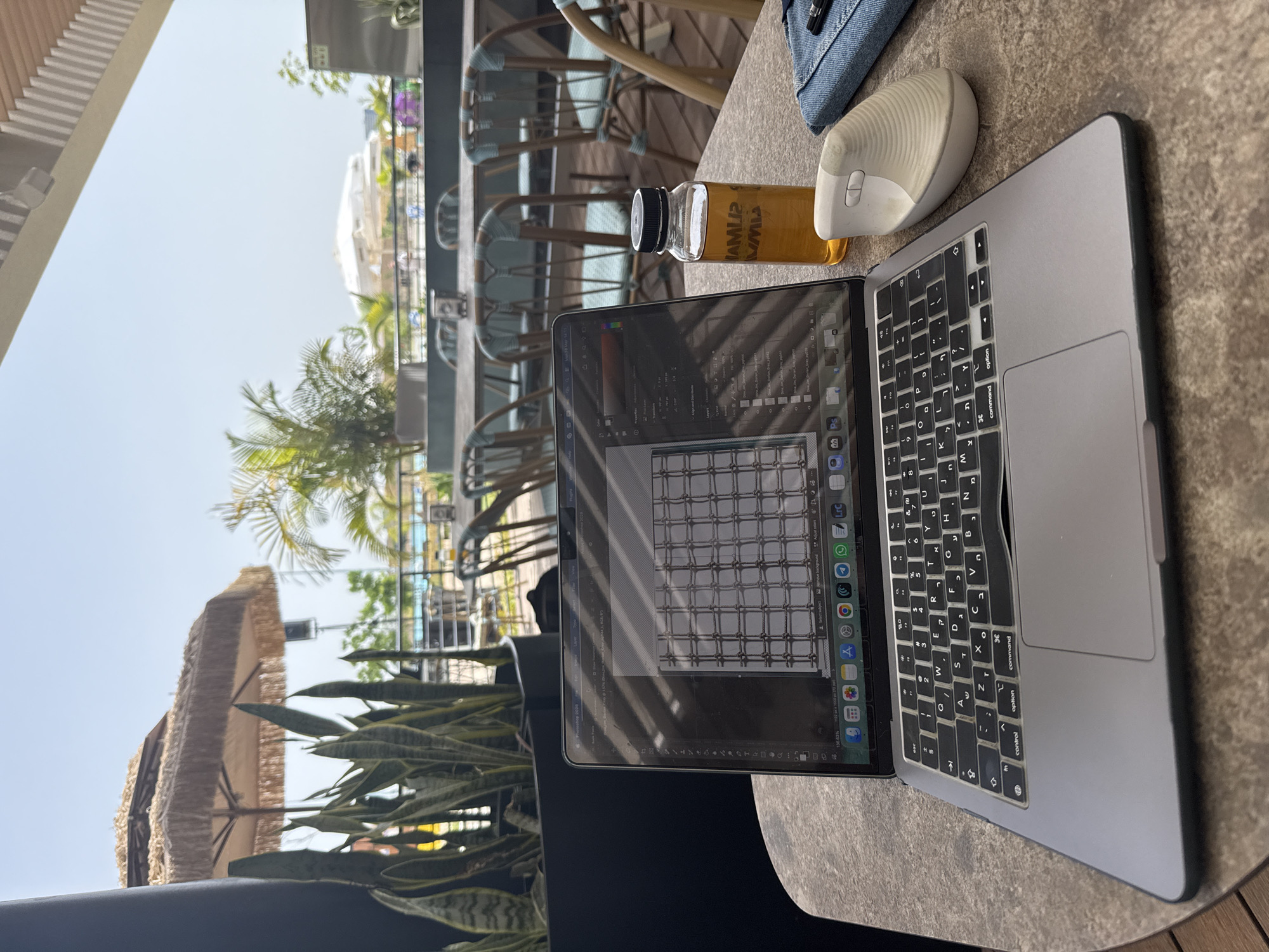 Here is a description of the image:
The image shows a laptop, a mouse, and a bottle of liquid on a table. The laptop is open, and the screen displays what appears to be a grid-like design within a graphic design program. A white ergonomic mouse sits next to the laptop. Next to the mouse is a bottle filled with a yellowish liquid, possibly juice or tea. The background shows an outdoor setting, possibly a cafe or patio, with chairs, umbrellas, and greenery visible. The image location appears to be in Tel Aviv, Israel, based on the objects in the view.