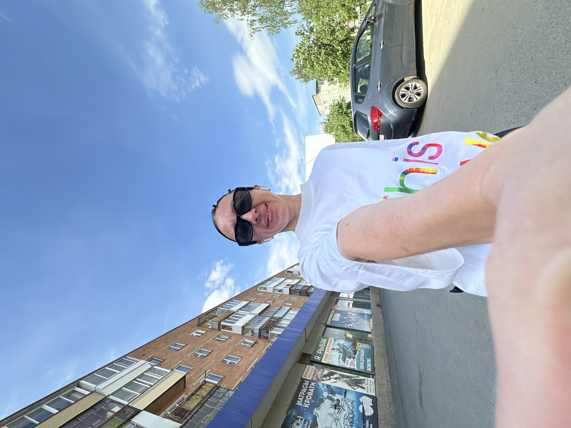 A woman wearing sunglasses and a white t-shirt with rainbow letters is taking a selfie on a street in Votkinsk, Russia. A multi-story brick apartment building with a blue awning over storefronts stands behind her. One of the signs on the storefronts reads "MATPACY KPOBATH." A dark gray car is parked on the street. The sky is blue with scattered white clouds.