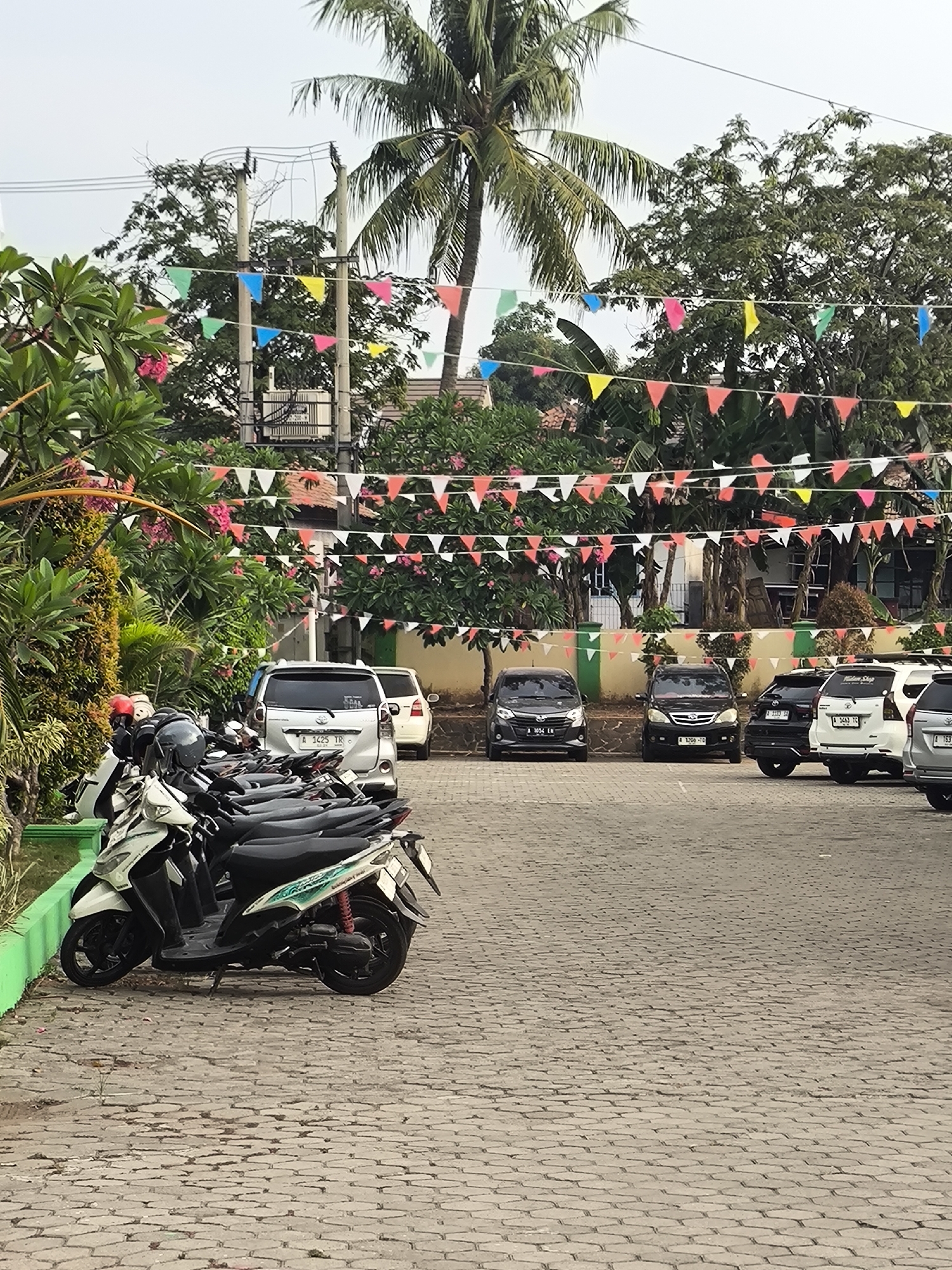 The image depicts an outdoor parking area in Cilegon, Indonesia. The ground is paved with interlocking gray bricks. In the foreground, a row of parked scooters is visible on the left side of the frame. Behind the scooters, several cars are parked in a lot, including a silver SUV and two black sedans. The background features lush green trees, including a prominent palm tree, and a building with an orange tiled roof. Colorful triangular flags are strung across the scene at various heights, adding a festive atmosphere. Electrical poles and overhead wires are also present. The lighting suggests it is daytime, possibly overcast.