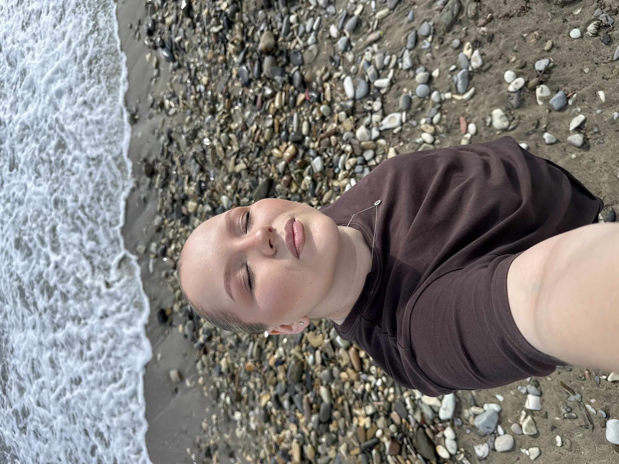 In an outdoor, high-angle, selfie-style photograph taken on a pebble beach near the city of Lermontovo, Russia, a young Caucasian woman is centered in the frame, her face turned upwards with eyes closed, and her lips slightly parted. She has blonde hair pulled back and is wearing dark brown clothing, accessorized with a small necklace. The background consists of a textured shoreline with numerous pebbles and a portion of the sea with gentle waves lapping at the edge. The lighting suggests it is daytime.
