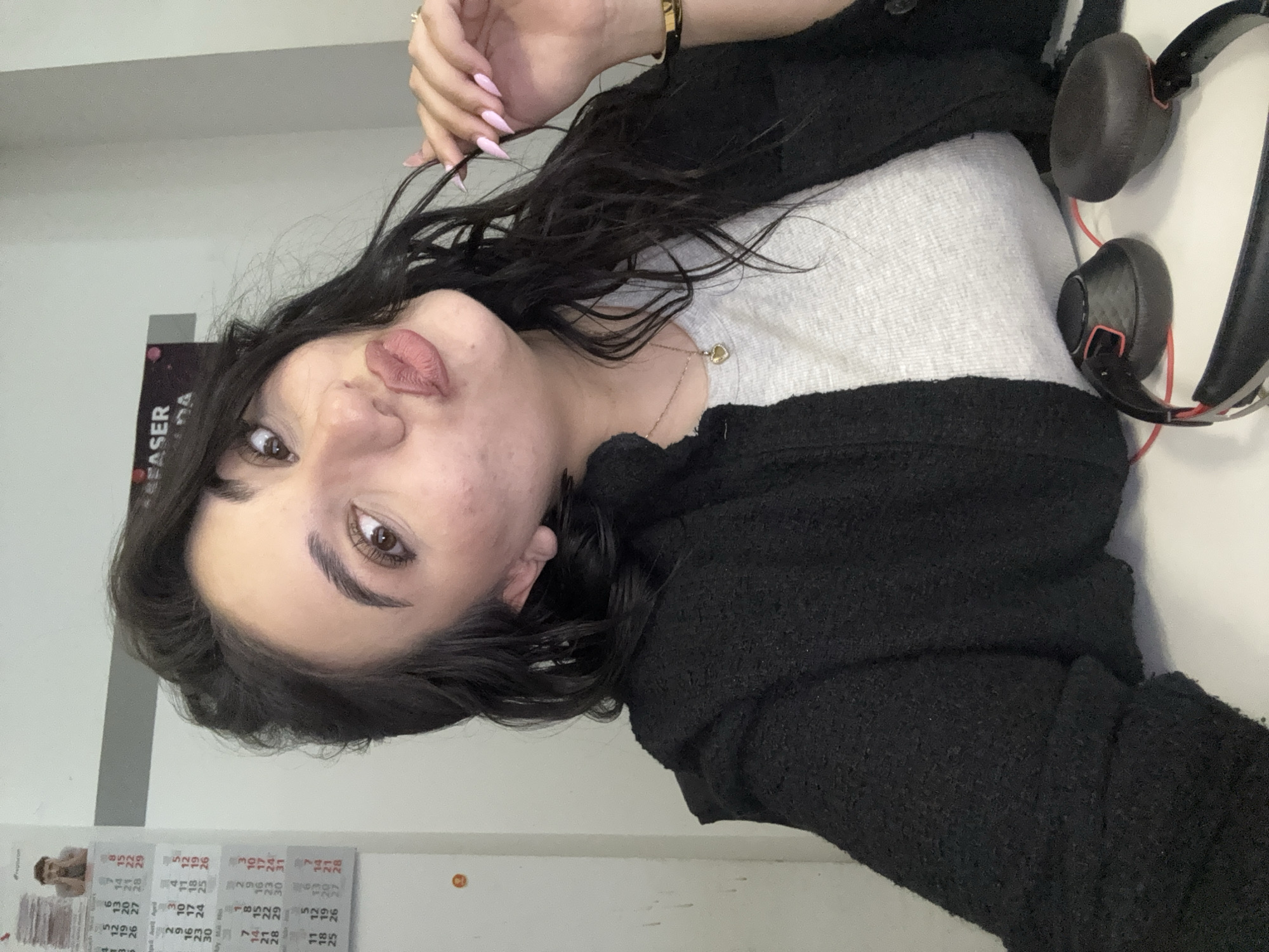 A woman is posing for a photo. She has dark hair, brown eyes, and light pink nails. She is wearing a black jacket over a light grey top, and a gold necklace. The woman has a ring on her finger and a gold bracelet on her wrist. A pair of headphones are resting on a white surface next to her. A calendar is visible in the background, but the location cannot be determined as Sofia, Bulgaria based on this image alone.