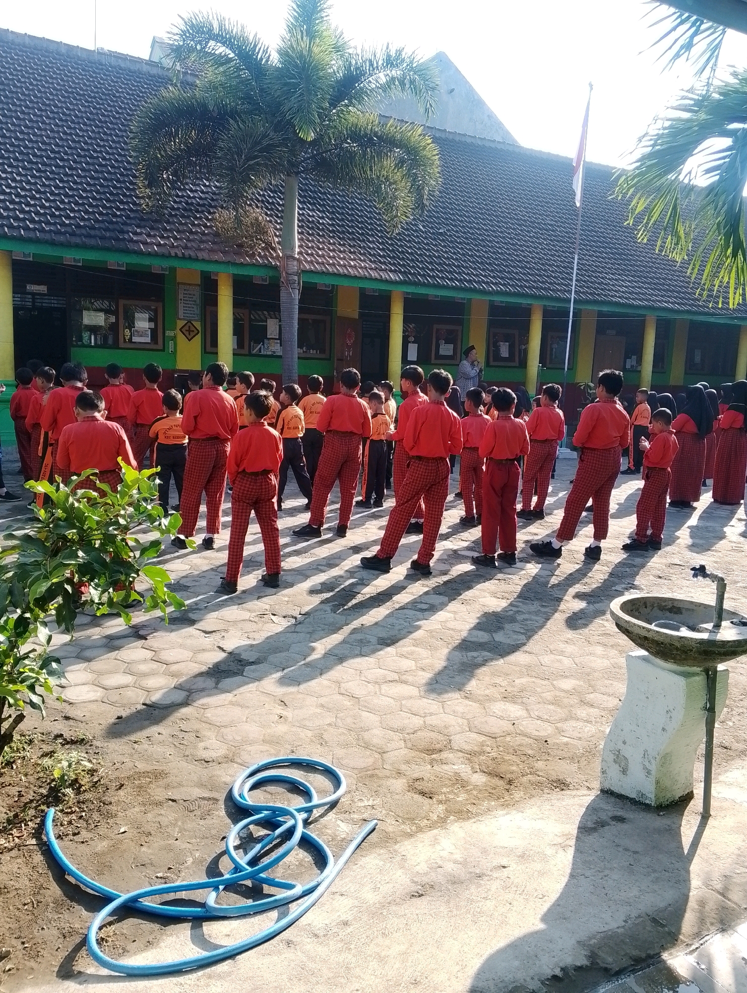 In front of a school building with a dark tiled roof and green and yellow trim, a group of children in red uniforms and red and black checkered pants stand in rows on a paved courtyard. The children are mostly facing away from the camera, with some looking towards a person standing in front of them. The school building has several windows and what appears to be posters or notices displayed. A flagpole with a red and white Indonesian flag is visible in the background. To the left of the children, there is a lush green bush. In the foreground, a blue garden hose is coiled on the ground. To the right, a concrete structure with a stone sink is present, casting a long shadow. The scene is illuminated by bright sunlight, creating distinct shadows across the courtyard. The location is identified as Papungan, Indonesia.