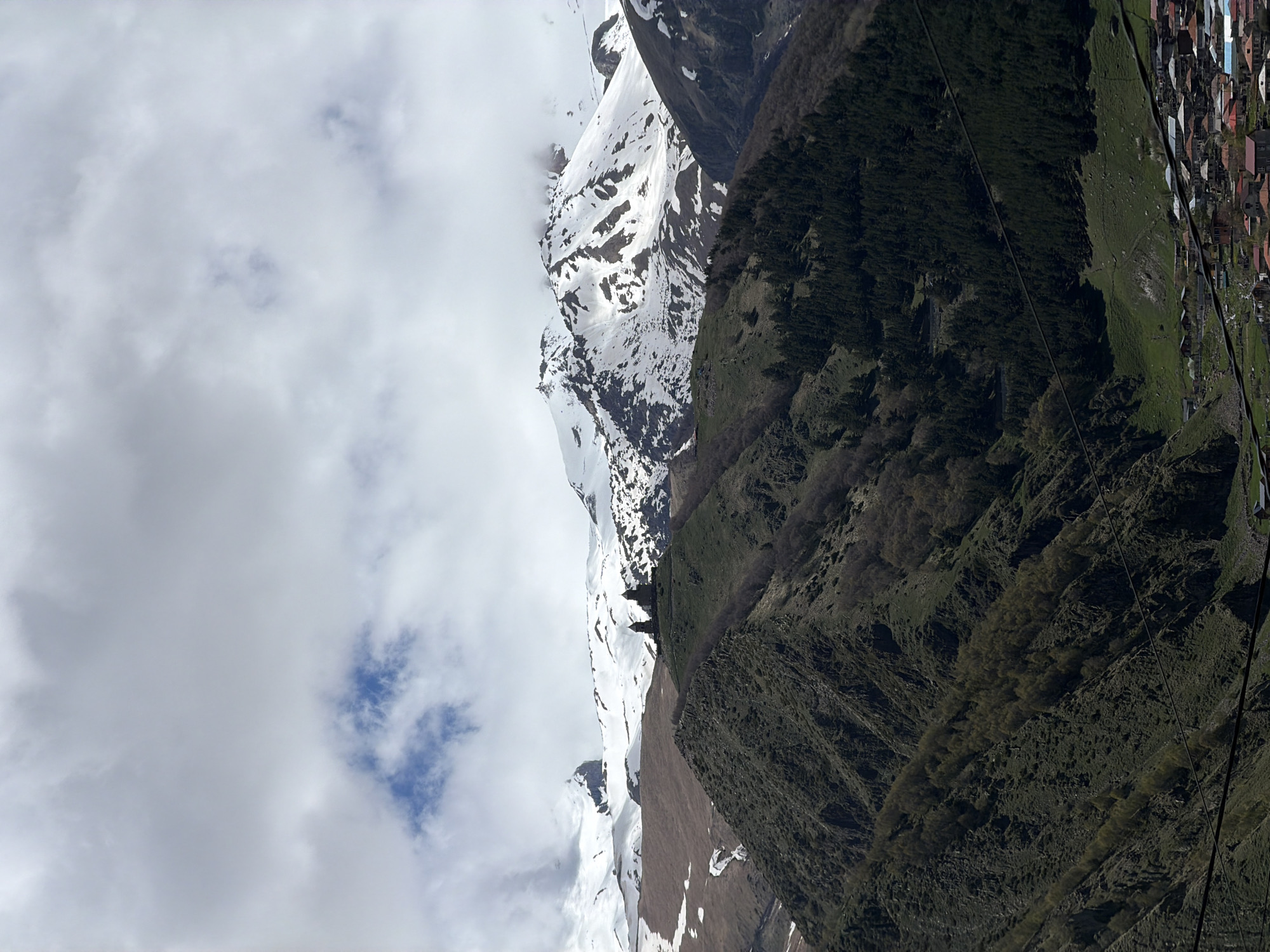 The image depicts a mountainous landscape in Stepantsminda, Georgia. On the left, snow-capped mountains are partially obscured by clouds. A steep, green mountainside occupies the right half of the image. A small town is visible on the upper-right edge. Power lines run along the right side of the mountain.