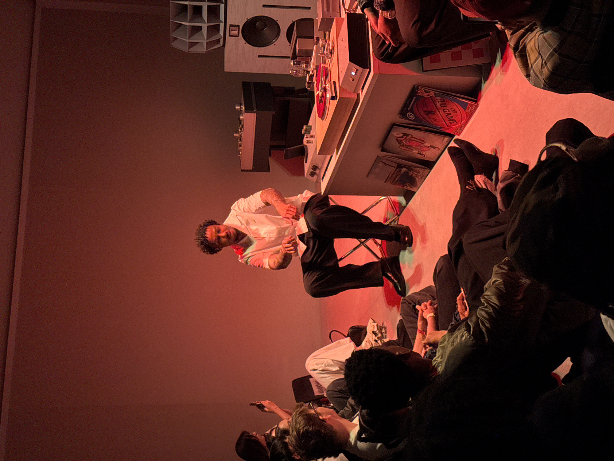 A man in a white shirt and black pants is performing in front of an audience in what appears to be a music venue. The man is sitting on a stool near a table with turntables and speakers. A group of people are watching the performance. The room is bathed in red light. The scene is potentially happening in London, United Kingdom but there is nothing in the image to verify the city.