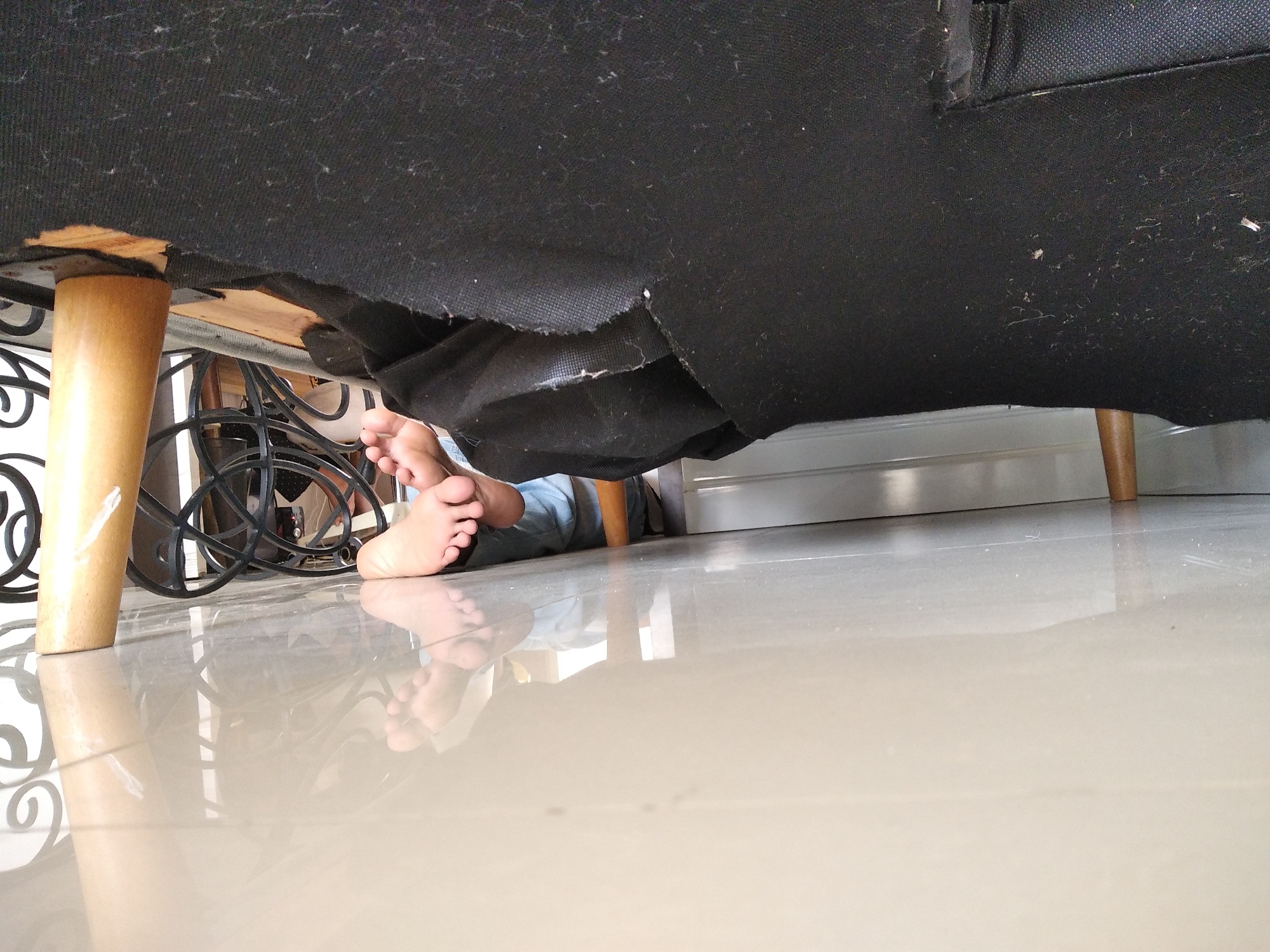 The image shows a low-angle view from under a dark-colored sofa or chair. A person's bare feet and lower legs are visible, positioned underneath the furniture. The feet appear to be resting on a light-colored tiled floor, which reflects the light. The sofa's black fabric appears somewhat worn or damaged in places, with exposed wooden supports and torn material. To the left, a wooden furniture leg is visible, and behind it, part of a black metal decorative scrollwork piece is seen. The environment suggests an indoor setting, likely a living room or similar space. The location in Kaliwates, Indonesia, is not visually indicated within the image itself.