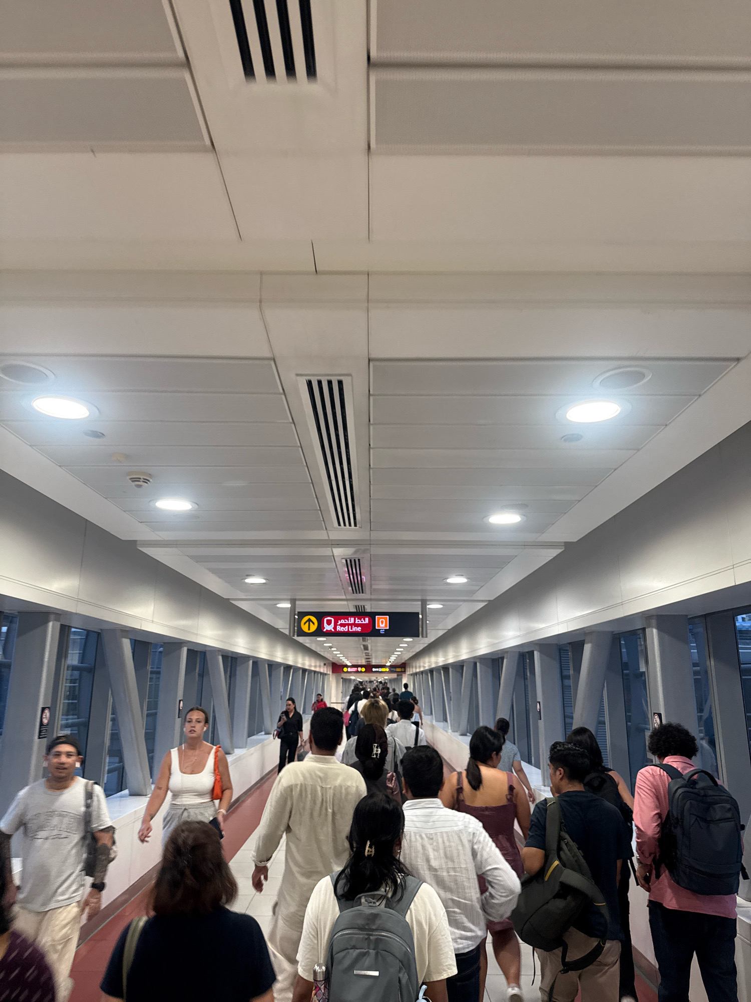This image depicts a pedestrian walkway in the city of Dubai, United Arab Emirates, likely within a modern public transportation hub such as a metro station. The walkway is enclosed by large glass panels, offering views of the city's architecture outside, which appears to be contemporary. The ceiling is composed of white modular panels with integrated lighting and air vents. A prominent directional sign with Arabic and English text, indicating "Red Line" for the metro, is visible. A diverse group of people, of various ethnicities and ages, are walking in both directions along the walkway, some carrying bags and backpacks. They are dressed in casual attire, reflecting the warm climate of Dubai. The overall atmosphere suggests a busy transit environment.