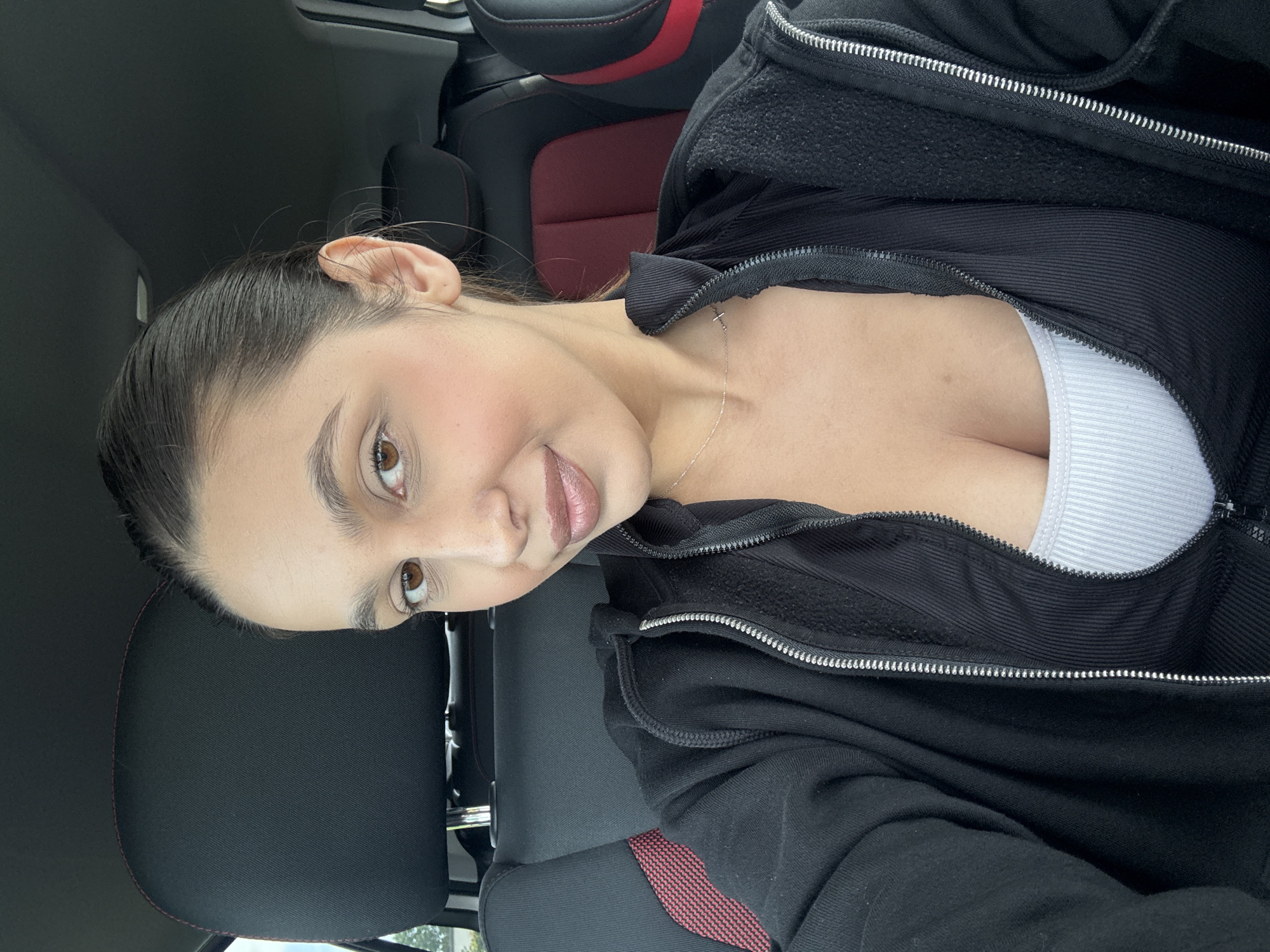 A female individual is depicted in a close-up, front-facing perspective, seated within the passenger compartment of a vehicle, in Wegberg, Germany. Her dark hair is pulled back from her face. She has brown eyes and appears to be wearing facial makeup, including a light application of blush on her cheeks and a nude-toned lip product. She is wearing a black, full-zip hooded sweatshirt, worn open, revealing a black ribbed top beneath, with a white ribbed tank top visible at the neckline. A thin, silver-colored necklace featuring a small cross pendant is visible around her neck. The individual is looking directly at the camera with a slight smile. The car interior features a black headrest with red stitching and black and red patterned seat upholstery. The ceiling of the vehicle is dark-colored.