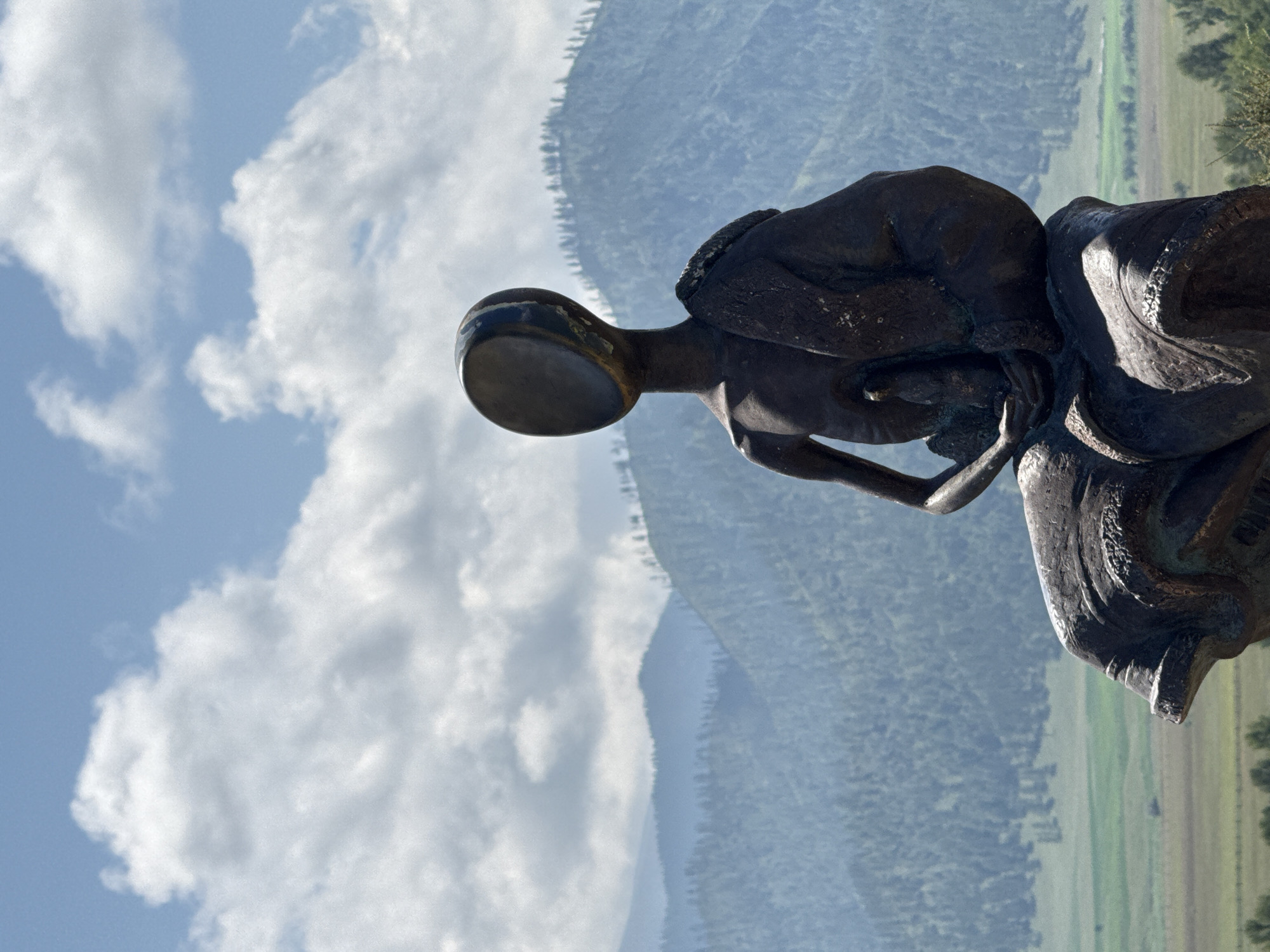 A dark bronze sculpture of a seated, faceless human figure is positioned in the foreground against a backdrop of layered, forested mountains and a bright blue sky with scattered white clouds. The sculpture depicts a figure with its head tilted back and its face an empty oval opening. The figure is draped in fabric that flows around its legs and shoulders, and its arms are held in front, cradling something small. The sculpture is weathered and appears to be made of metal, with visible patina and texture. The surrounding environment suggests an outdoor setting, likely a park or scenic overlook, with a valley visible in the distance between the sculpture and the mountains. The location is identified as Bichiktu-Boom, Russia.