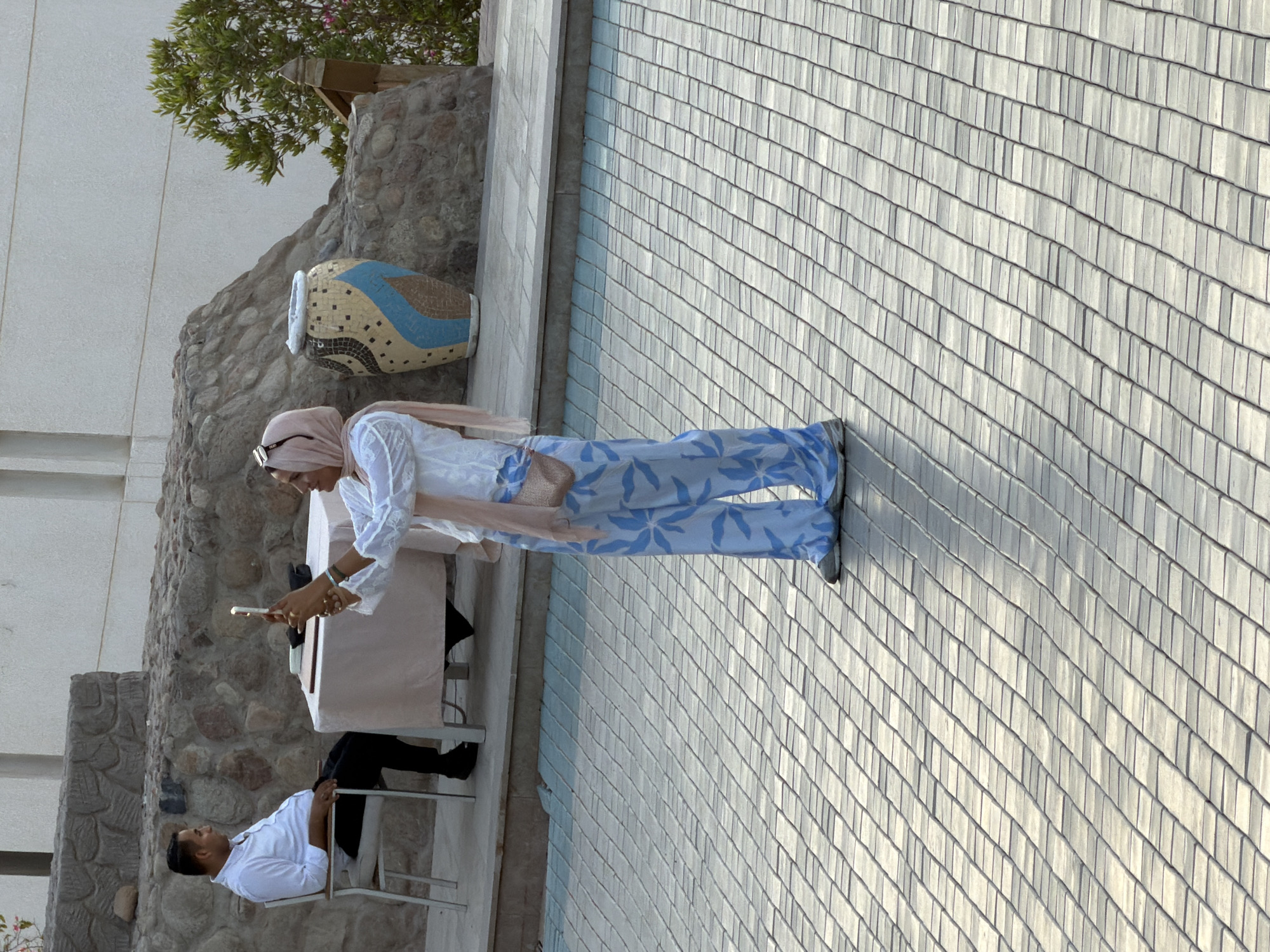 In Sharm El Sheikh, Egypt, a woman wearing a light-colored hijab and a white tunic with loose blue patterned pants stands on a paved walkway, holding a smartphone to take a picture. She is facing a man seated in a white chair at a table covered with a light pink cloth. The man is wearing a white shirt and black pants and is looking away from the woman. Behind the man and the table, there is a low stone wall. To the right of the wall, a large decorative ceramic vase with geometric patterns is visible. The environment appears to be an outdoor area, possibly a resort or public space, with natural rock formations and some greenery in the background. The paving stones in the foreground are laid in a diagonal pattern.
