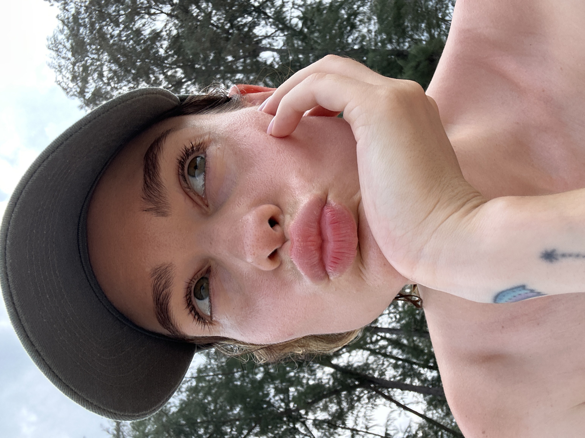 A fair-skinned person with light brown hair, wearing a grey cap, is posing outdoors with pursed lips. The person has a tattoo on their upper chest/shoulder. Trees and sky are visible in the background. Location information is not discernible from the image.