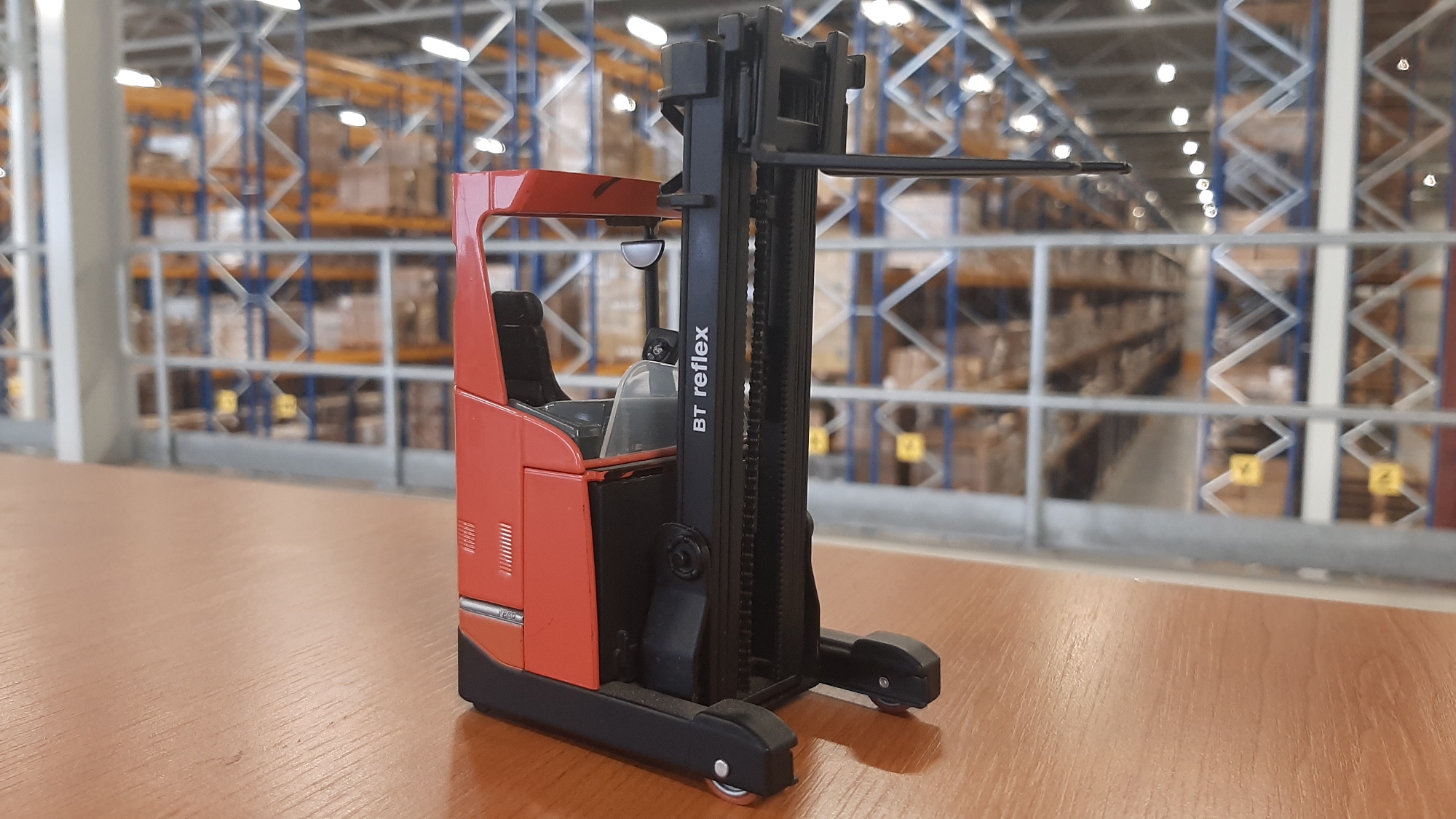 A close-up, low-angle shot captures a red and black toy forklift with the text "BT reflex" on its mast, positioned on a wooden surface. In the background, a blurred warehouse setting is visible with rows of shelves filled with boxes under bright, diffused lighting. The scene is static and does not depict any specific location in the city of Hellevoetsluis, Netherlands. There are no people or actions taking place in the image.