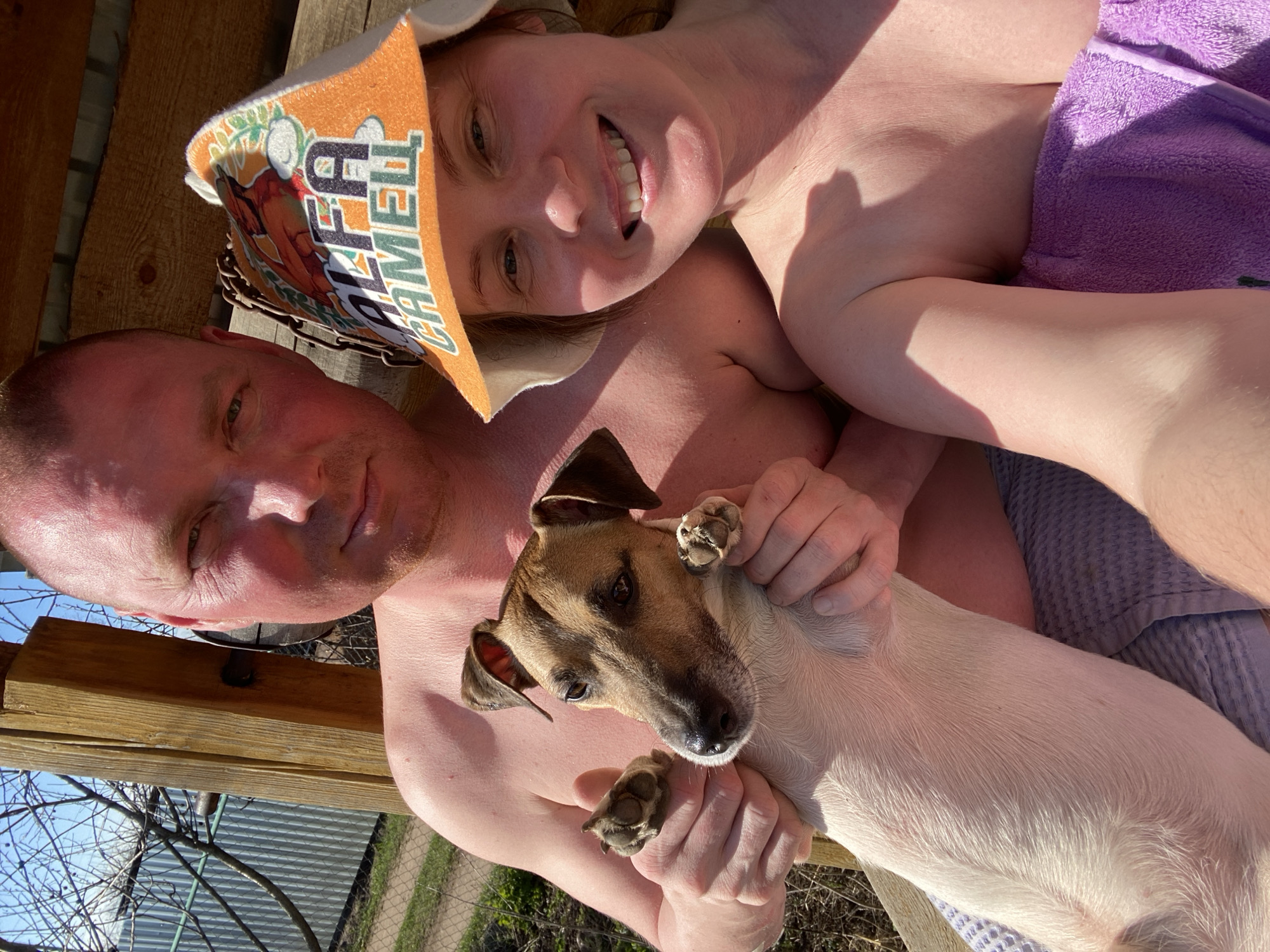 A sunny, close-up selfie shows a middle-aged Caucasian couple with a tan and a tan and brown dog. The man on the left has short, reddish hair and is looking into the camera. The woman on the top has reddish hair and is smiling. She is wearing an orange hat with the word "CAMEL" printed on it. The couple is holding a small dog. The dog has light brown fur with darker brown patches on its head and ears. The couple is sitting on what appears to be a wooden bench or deck. In the background, there is foliage and glimpses of a fence and trees, suggesting the photo was taken outdoors, possibly in a backyard or garden. The location is Chaikovsky, Russia.