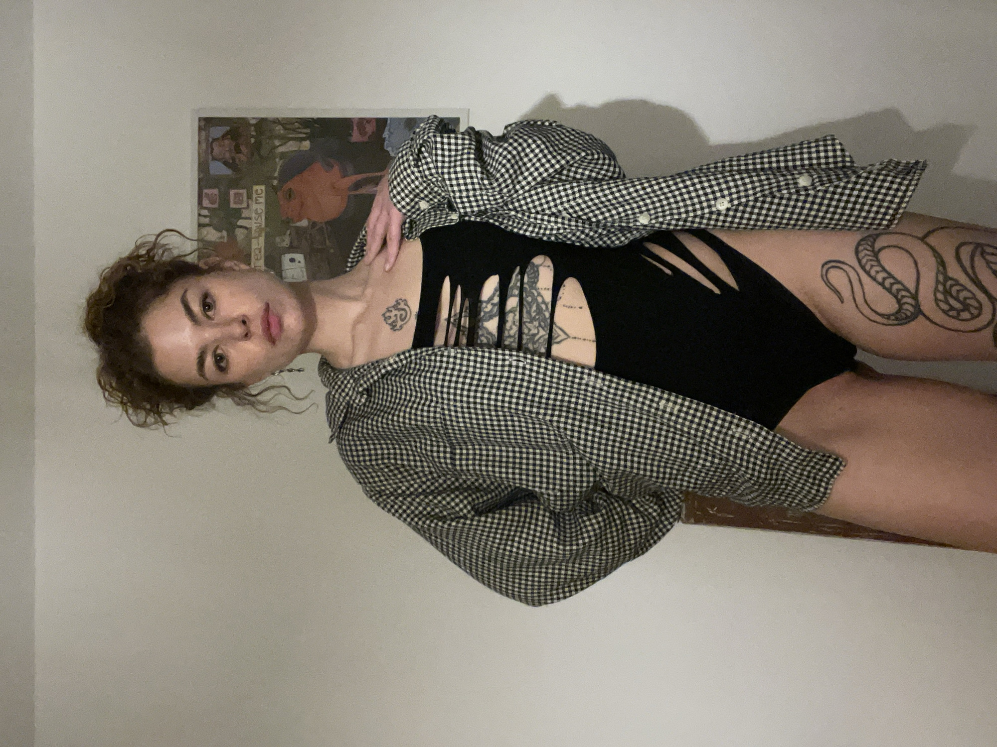 A woman is lying down in front of a white wall. She is wearing a black bodysuit with vertical cutouts down the front, and a black and white checkered shirt. She has curly brown hair and tattoos on her neck, chest, and thigh. Behind her is a framed poster. The location is not specified or discernible in the image.