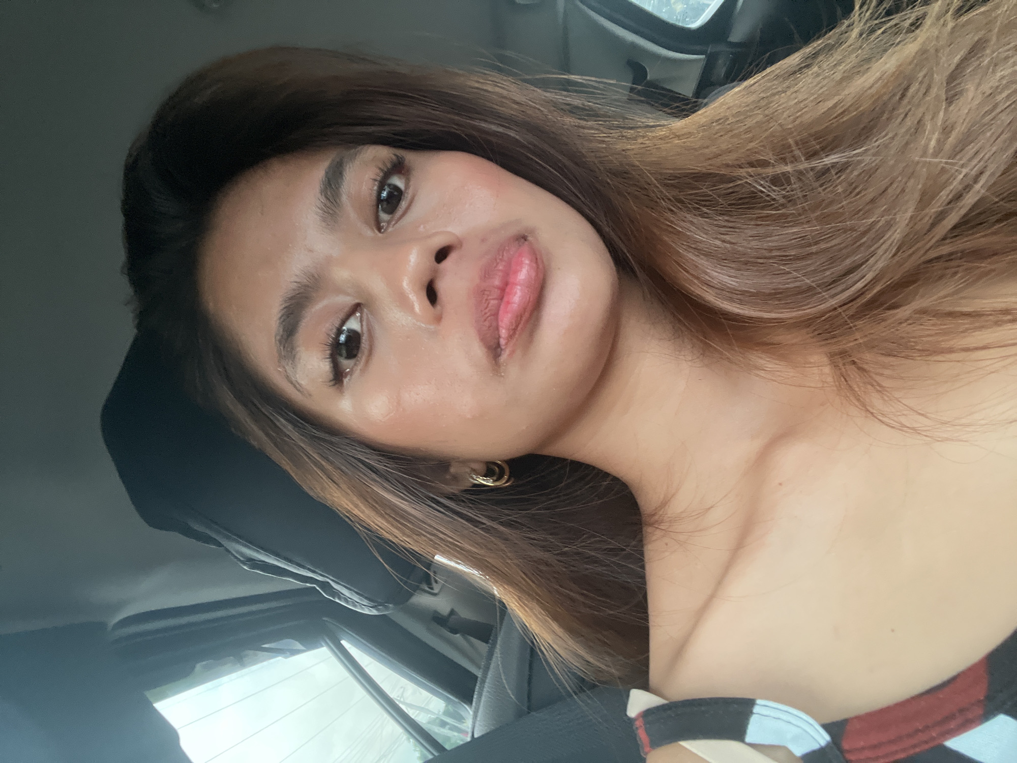 A young woman with brown hair and hoop earrings is sitting in the passenger seat of a car. She is looking at the camera with her lips slightly parted. The interior of the car is visible, including the headrest and the window. The scene is lit by natural light. The location is Panabo City, Philippines.