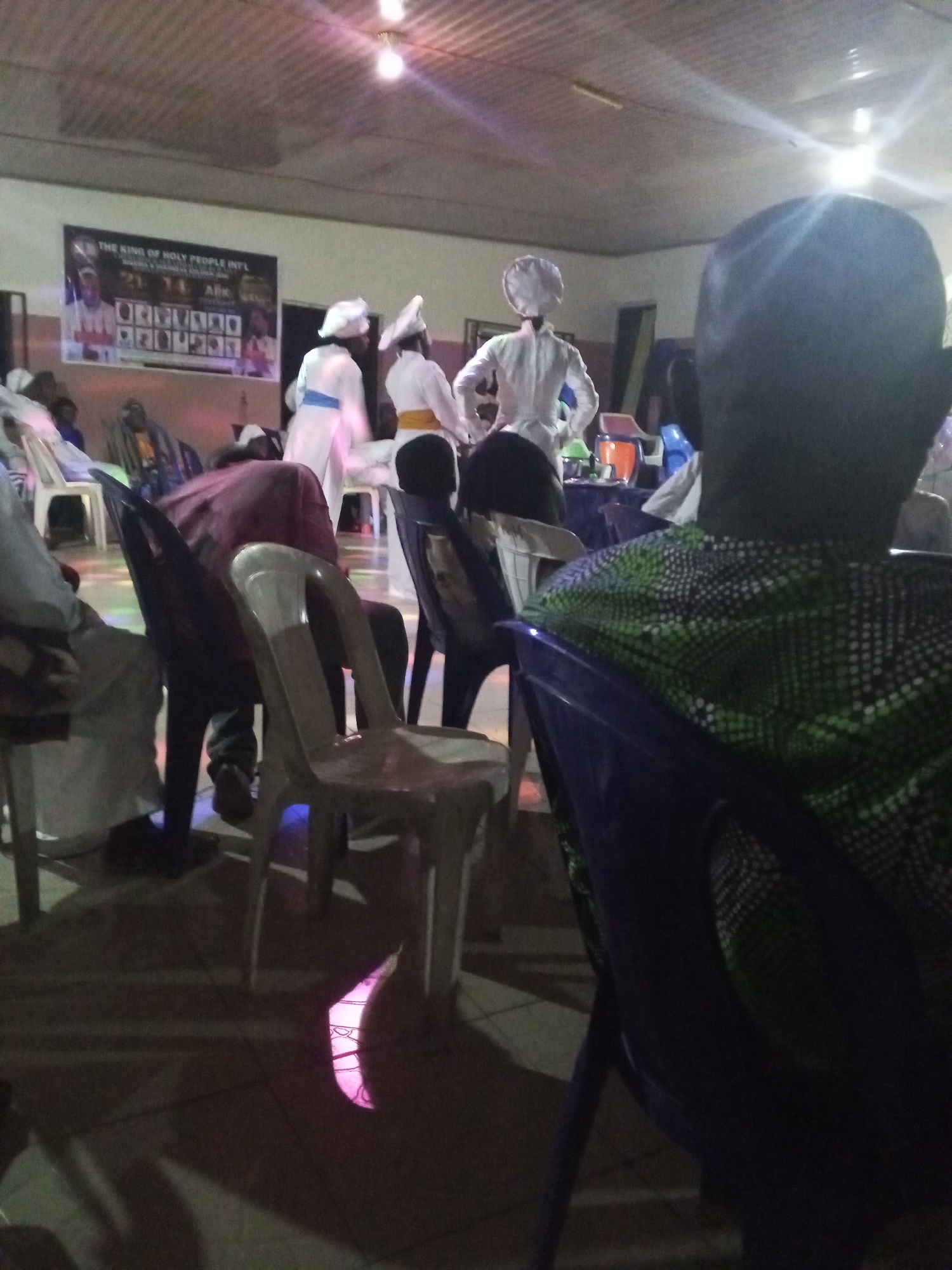The image captures an indoor scene, likely a religious gathering in Lokoja, Nigeria. Several people are seated on plastic chairs, facing a stage or platform where individuals dressed in white uniforms and head coverings are present. A banner with the words "THE KING OF HOLY PEOPLE INT'L" is visible in the background. The lighting is dim, illuminated by a few overhead lights. The floor appears to be tiled. In the foreground, the back of a person wearing a green and black patterned shirt is visible, partially obscuring the view of the rest of the room. The overall atmosphere appears to be focused on the stage where the people in white are standing.