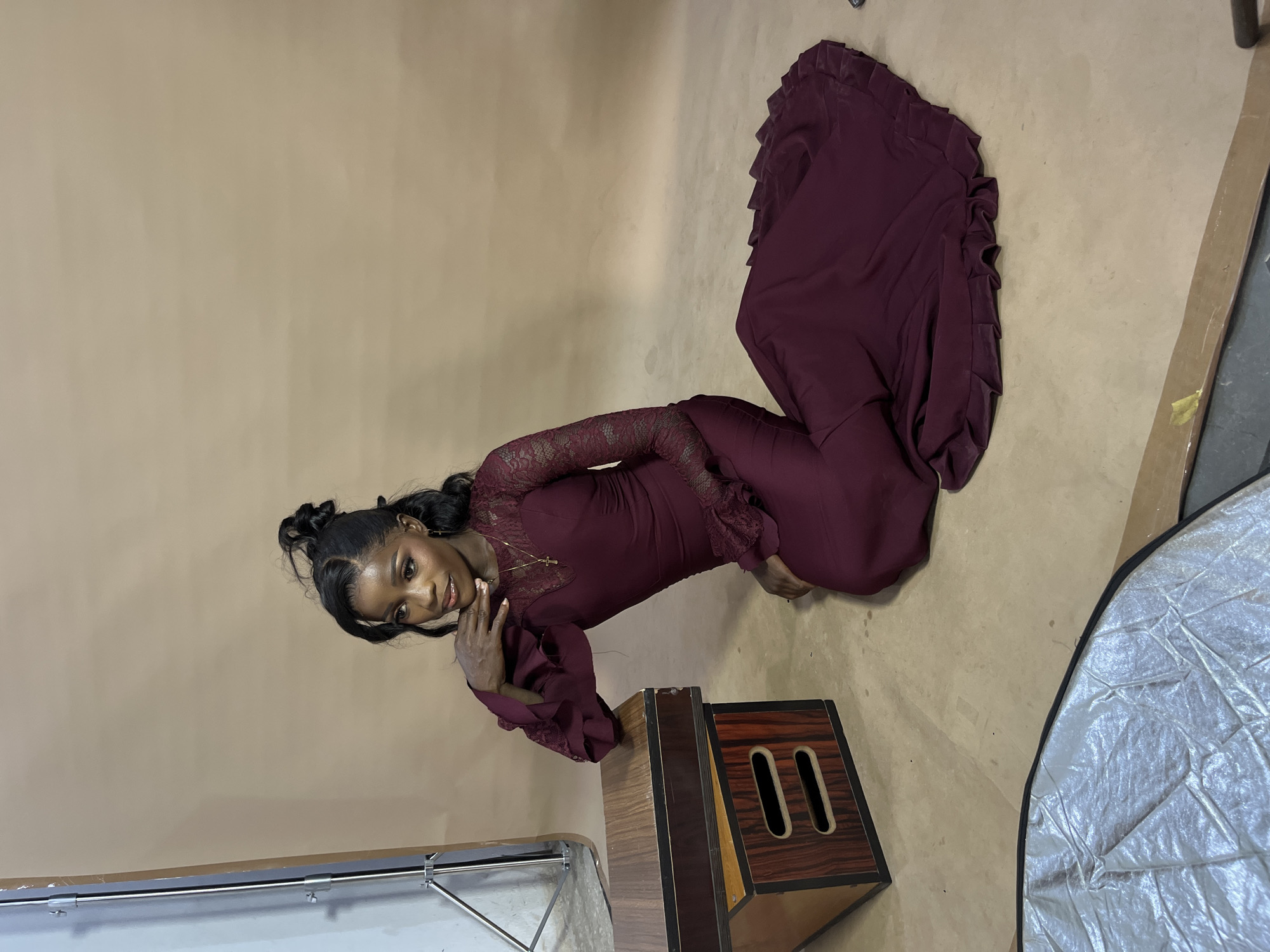 A young Black woman is posed on the floor in a studio setting, wearing a long, deep red, floor-length gown with lace sleeves and bodice. Her hair is styled in a high bun with loose tendrils framing her face. She is resting her chin on her hand, looking to the left of the frame. In front of her, to the left, are two wooden boxes stacked on top of each other, with the top box appearing to be slightly angled. The floor is a light brown, textured surface, and the background is a solid, muted tan. There is also a reflective silver object, possibly a piece of photography equipment, visible in the lower left corner of the frame. The overall scene suggests a professional photoshoot. There is no specific indication of the city of Gura Top, Nigeria.