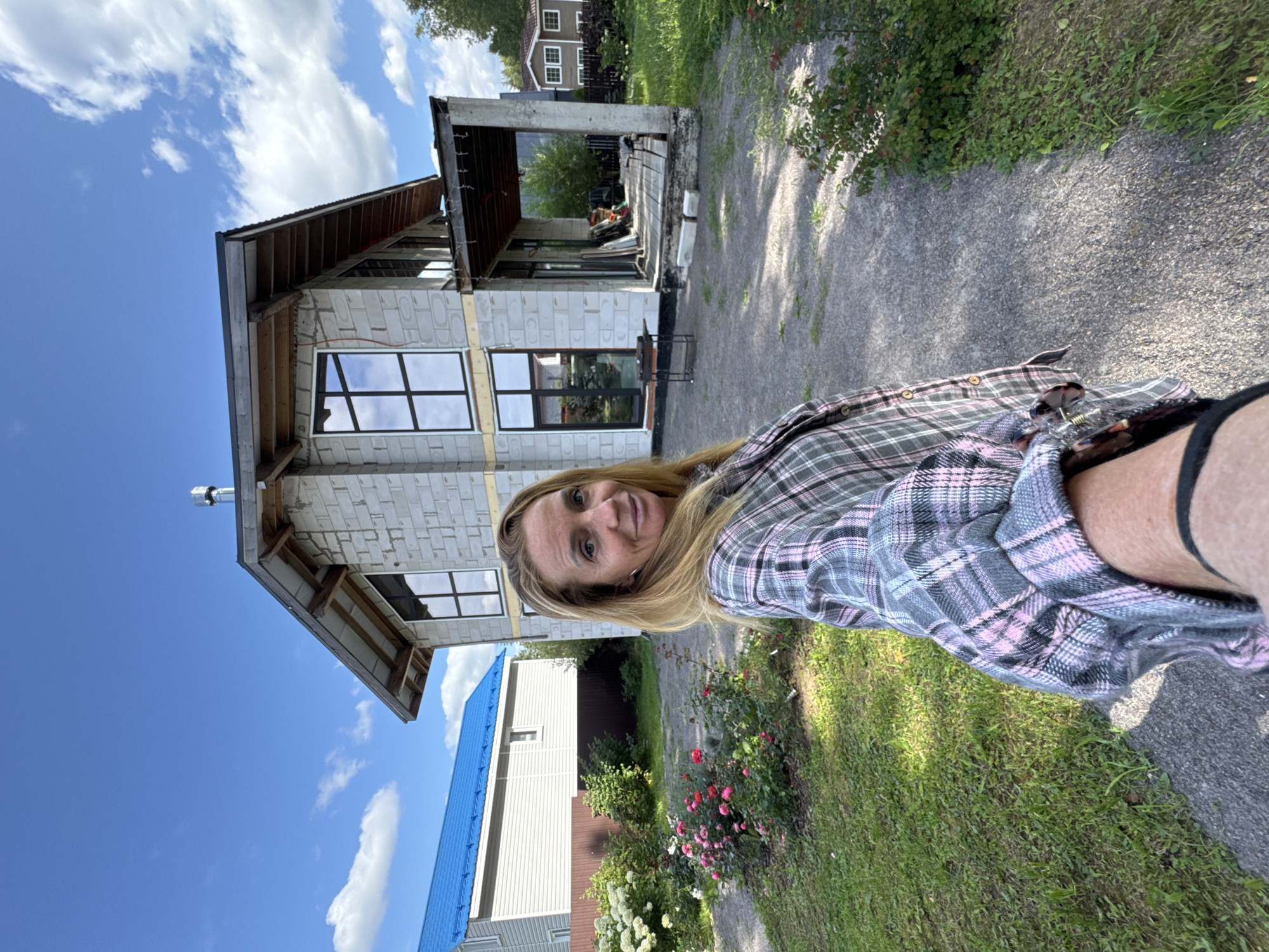 In Steklyannyi, Russia, a woman with blonde hair is taking a selfie in front of a house under construction. The house is made of concrete blocks and has large windows. The woman is wearing a plaid shirt. The yard is unkempt, with a gravel path leading to the house and a garden with roses. In the background, another house with a blue roof is visible. The sky is blue with scattered clouds.